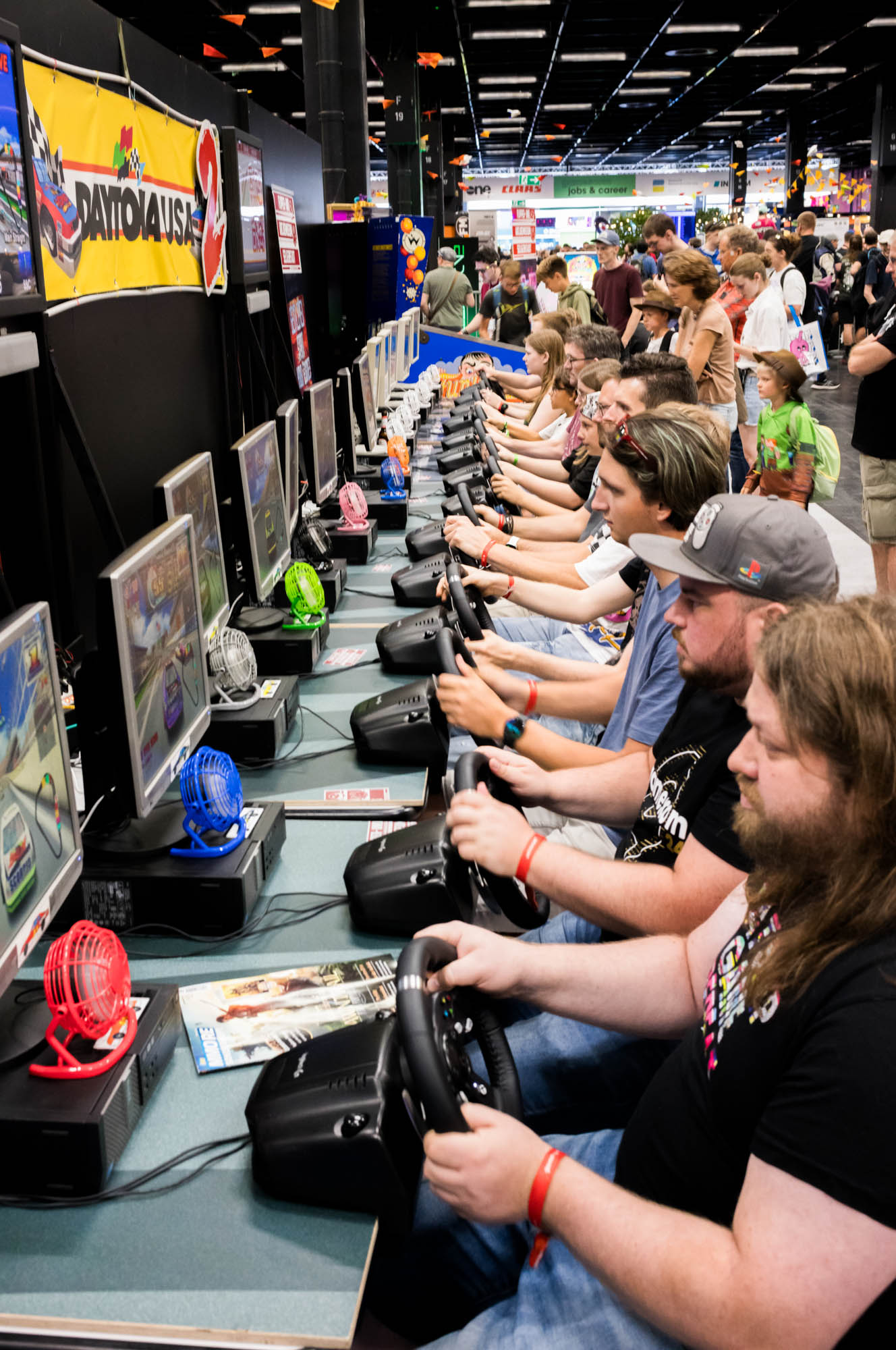 People playing racing games at gaming expo, using steering wheel controllers in a row of arcade setups.