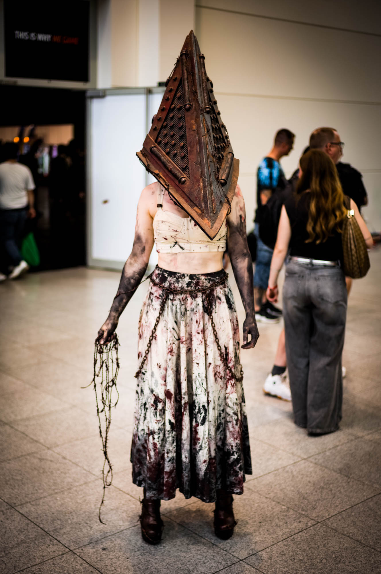 Person in horror-themed cosplay with pyramid helmet at a convention, holding chains, back view.