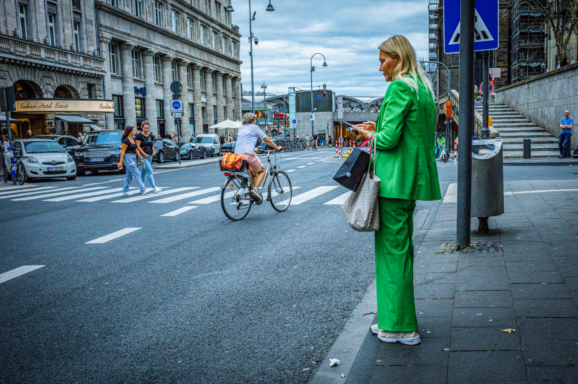 Woman in green suit stands by crosswalk, holding shopping bags, while cyclist rides by in a busy city street.
