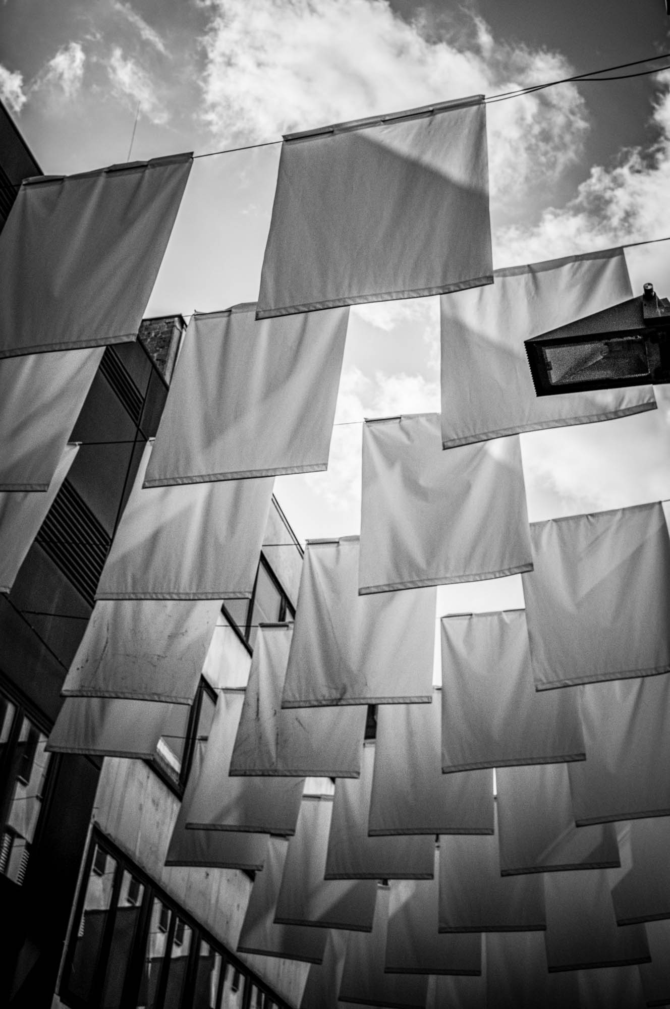 Monochrome view of white flags hanging in an alley against a cloudy sky, emphasizing geometric shapes and lighting.