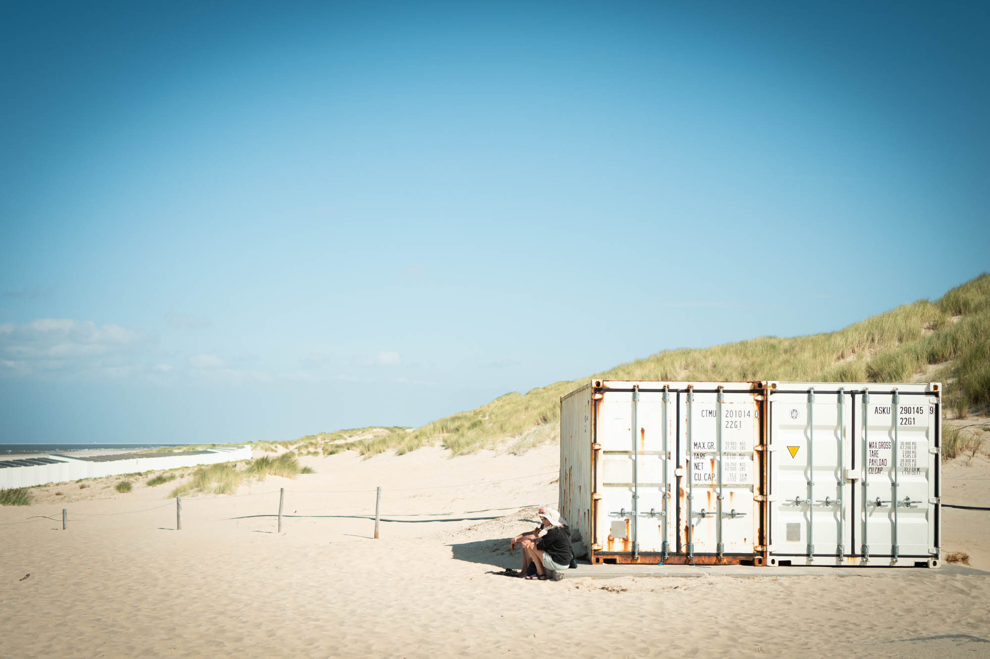 Person sitting by a shipping container on a sandy beach under clear blue sky.