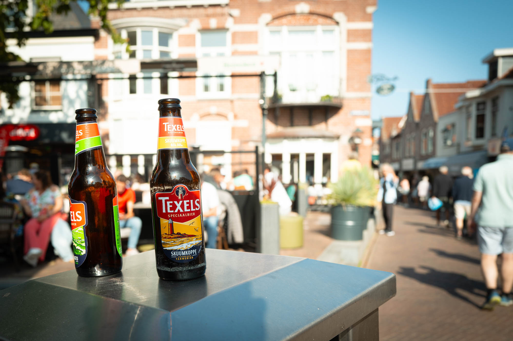 Two Texels beer bottles in a sunny street setting with people and buildings in the background.