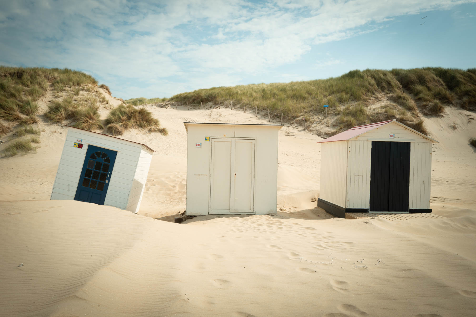 Beach huts on a sandy shore with grassy dunes under a blue sky.