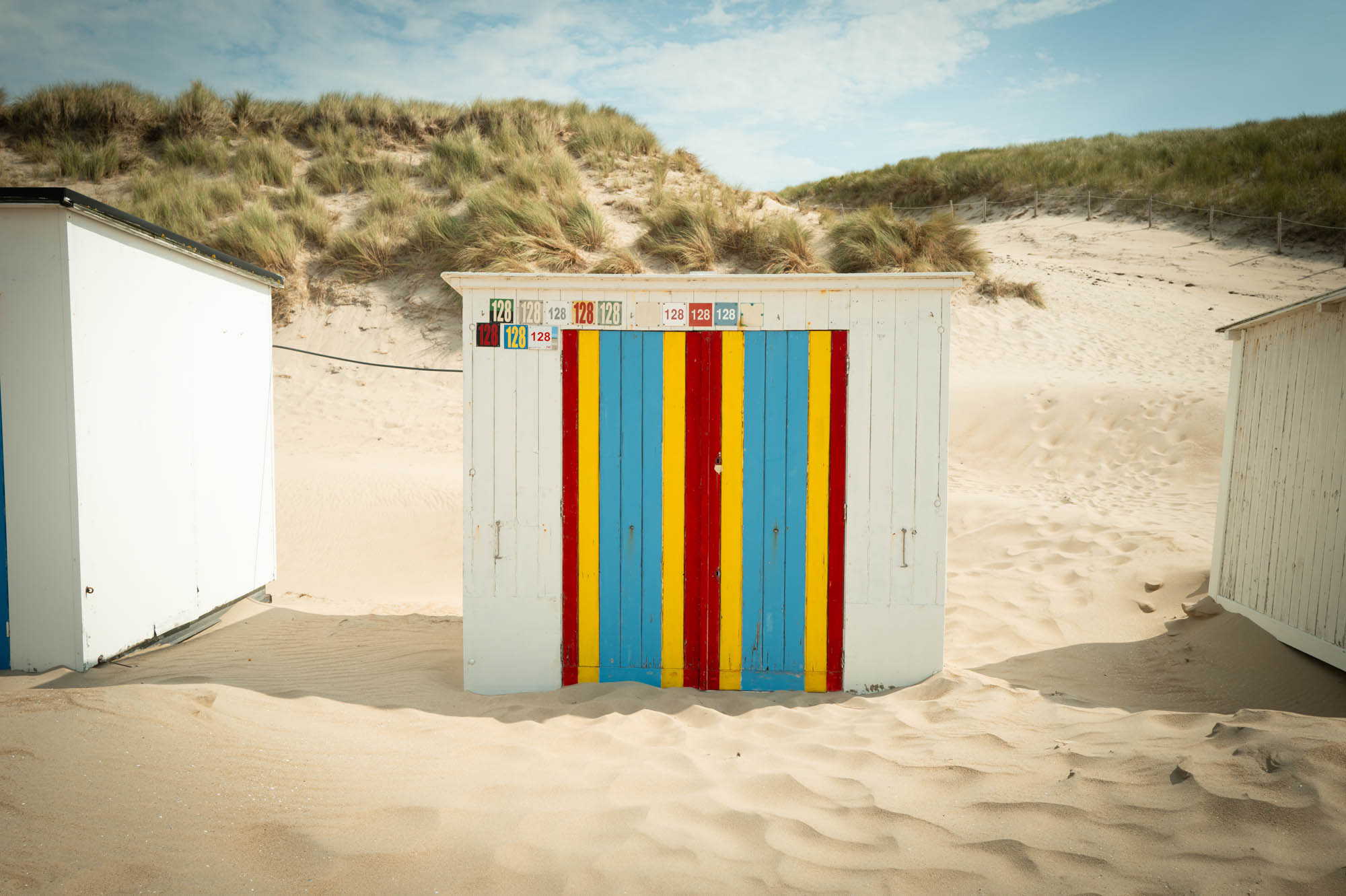 Colorful beach hut with blue, red, and yellow stripes on a sandy dune under a clear sky.