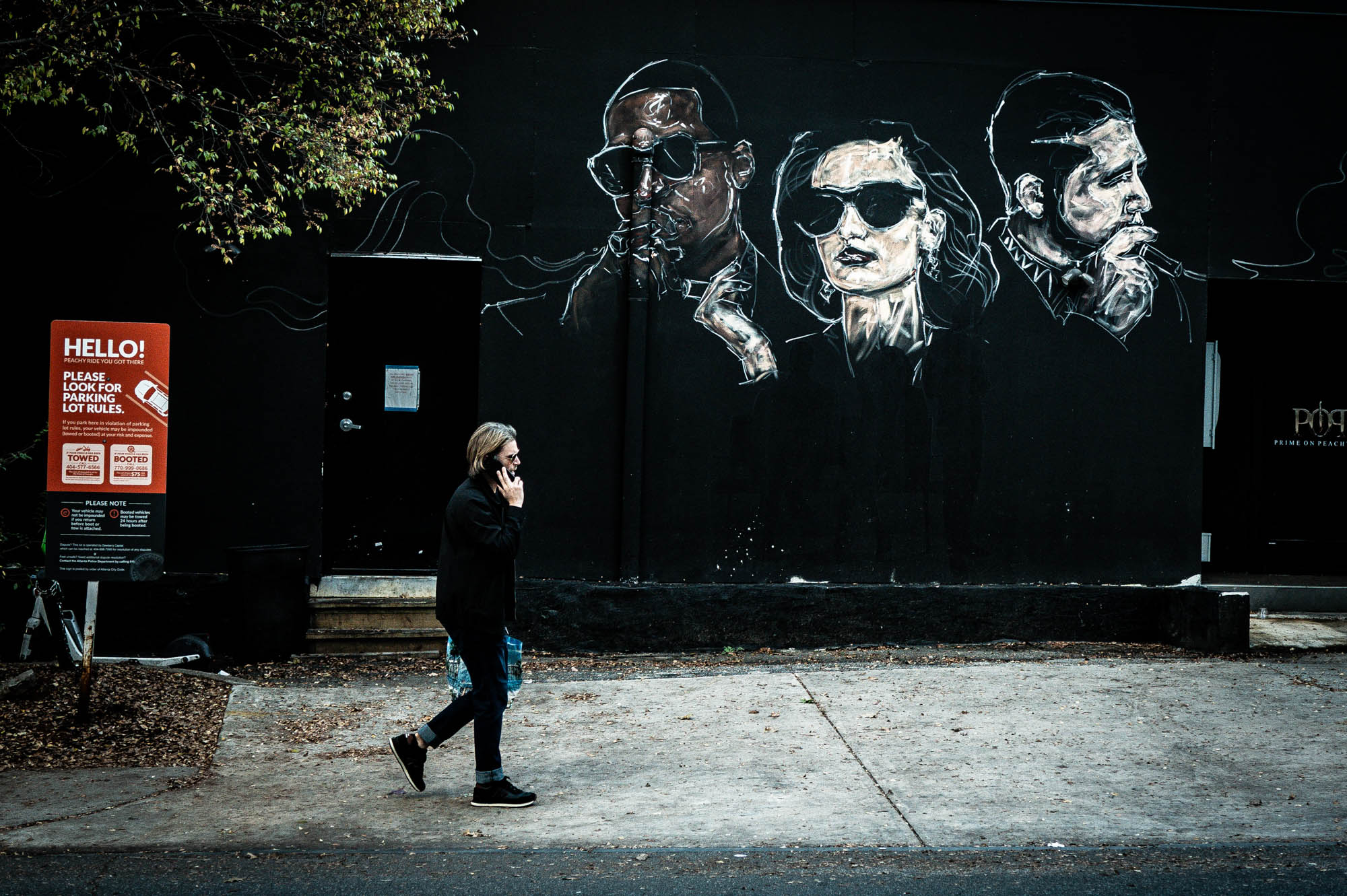 Person walking past street art mural with three faces wearing sunglasses, next to a parking rules sign.
