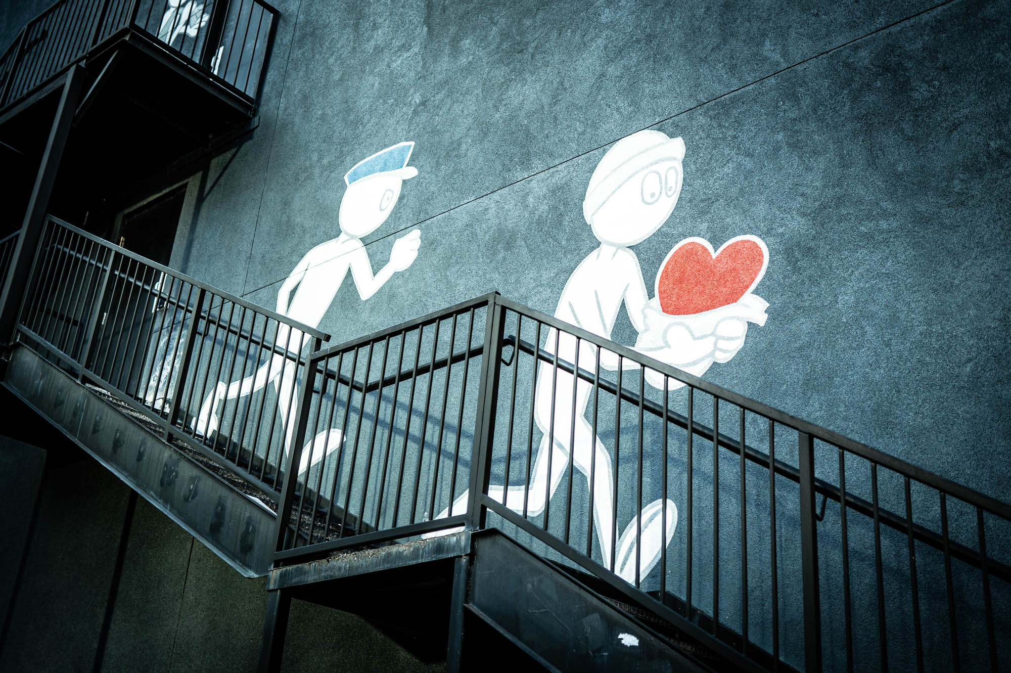 Street art on a wall depicting a figure chasing another with a heart, viewed from a metal staircase.