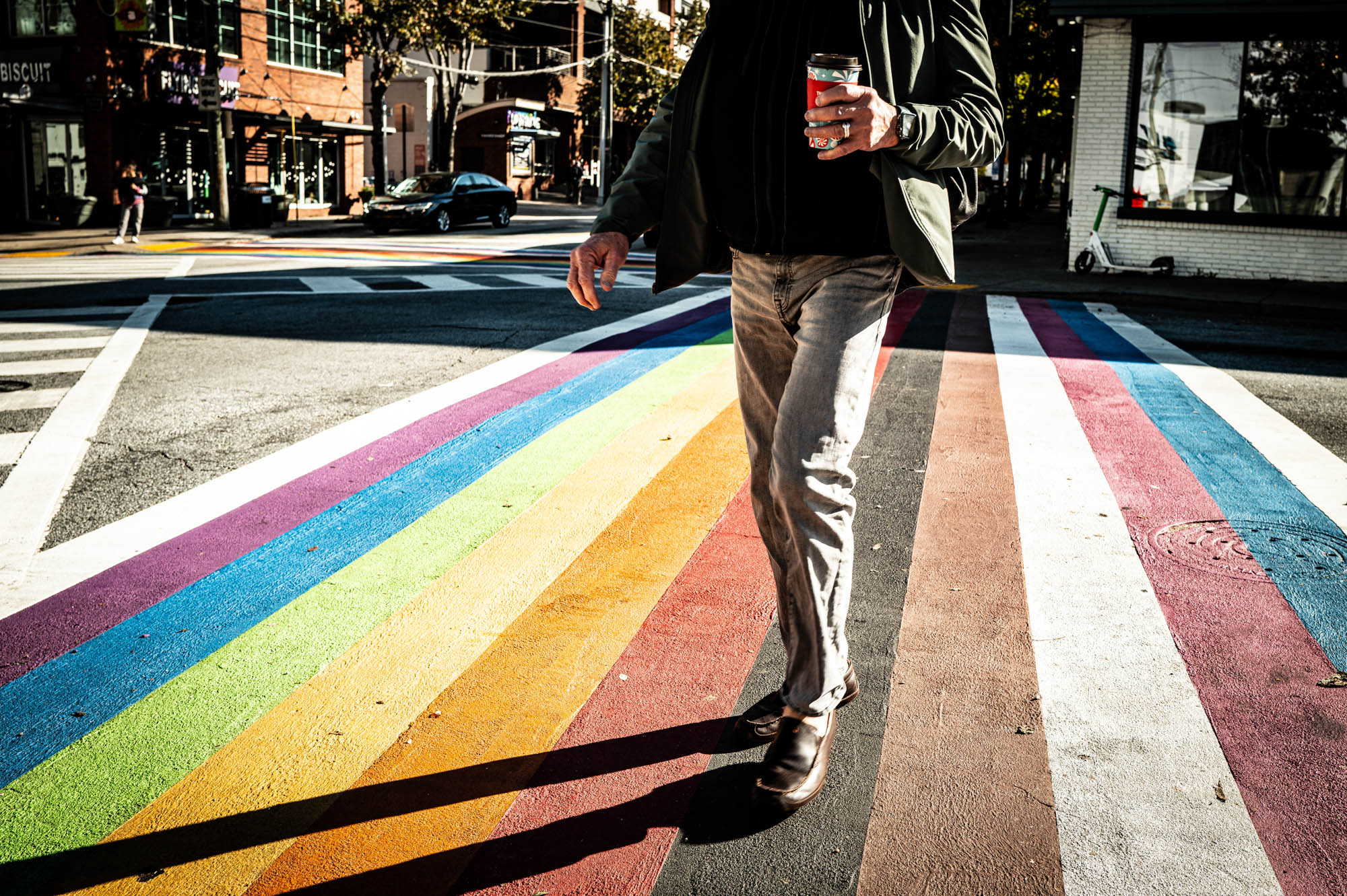 Person walking on a vibrant rainbow crosswalk, holding a red cup, in a sunny urban street scene.
