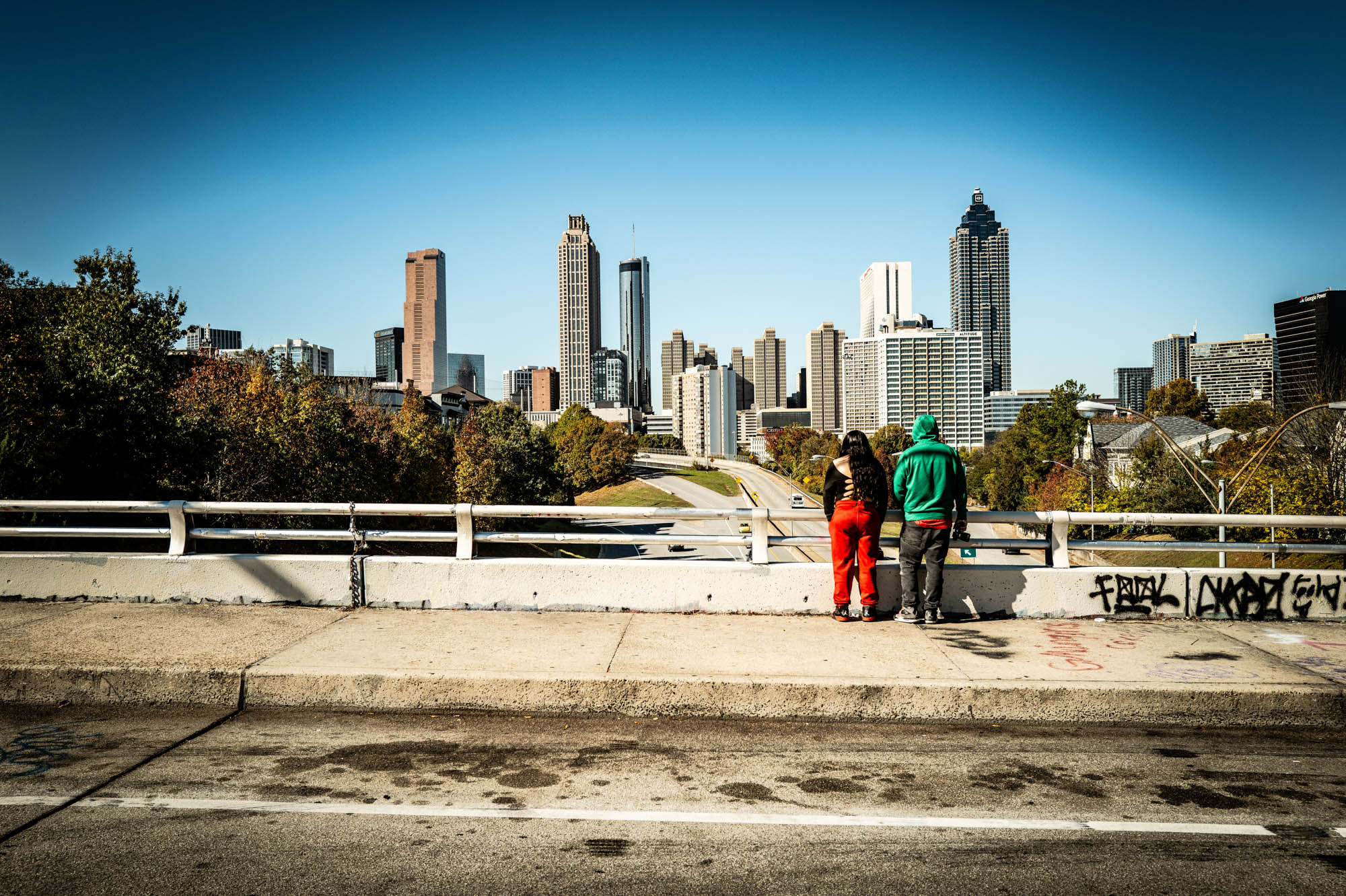 Couple admires Atlanta skyline from a bridge, surrounded by clear blue sky and vibrant autumn foliage.