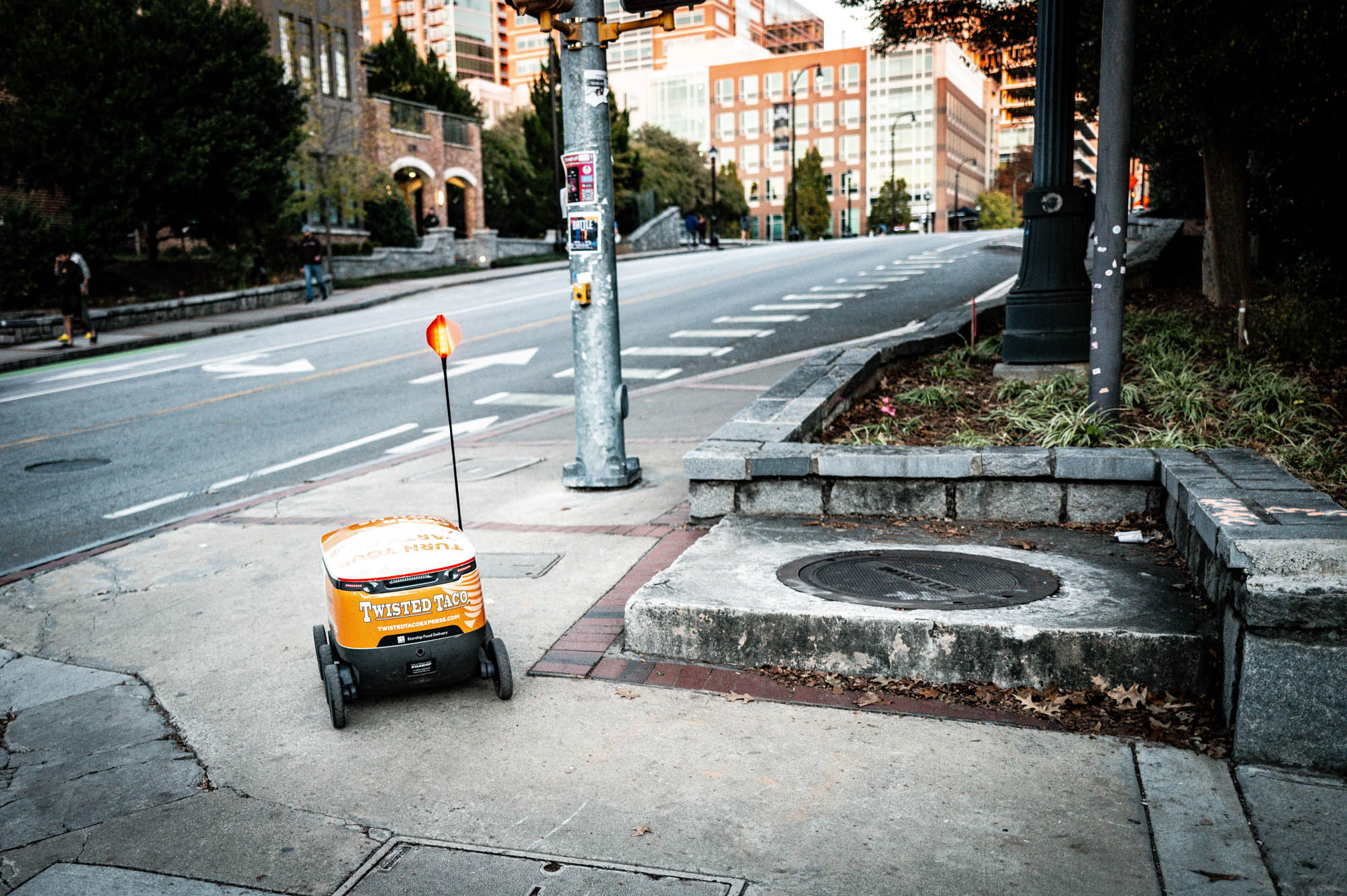 Delivery robot on an urban sidewalk, branded Twisted Taco, near a city street with buildings and greenery.