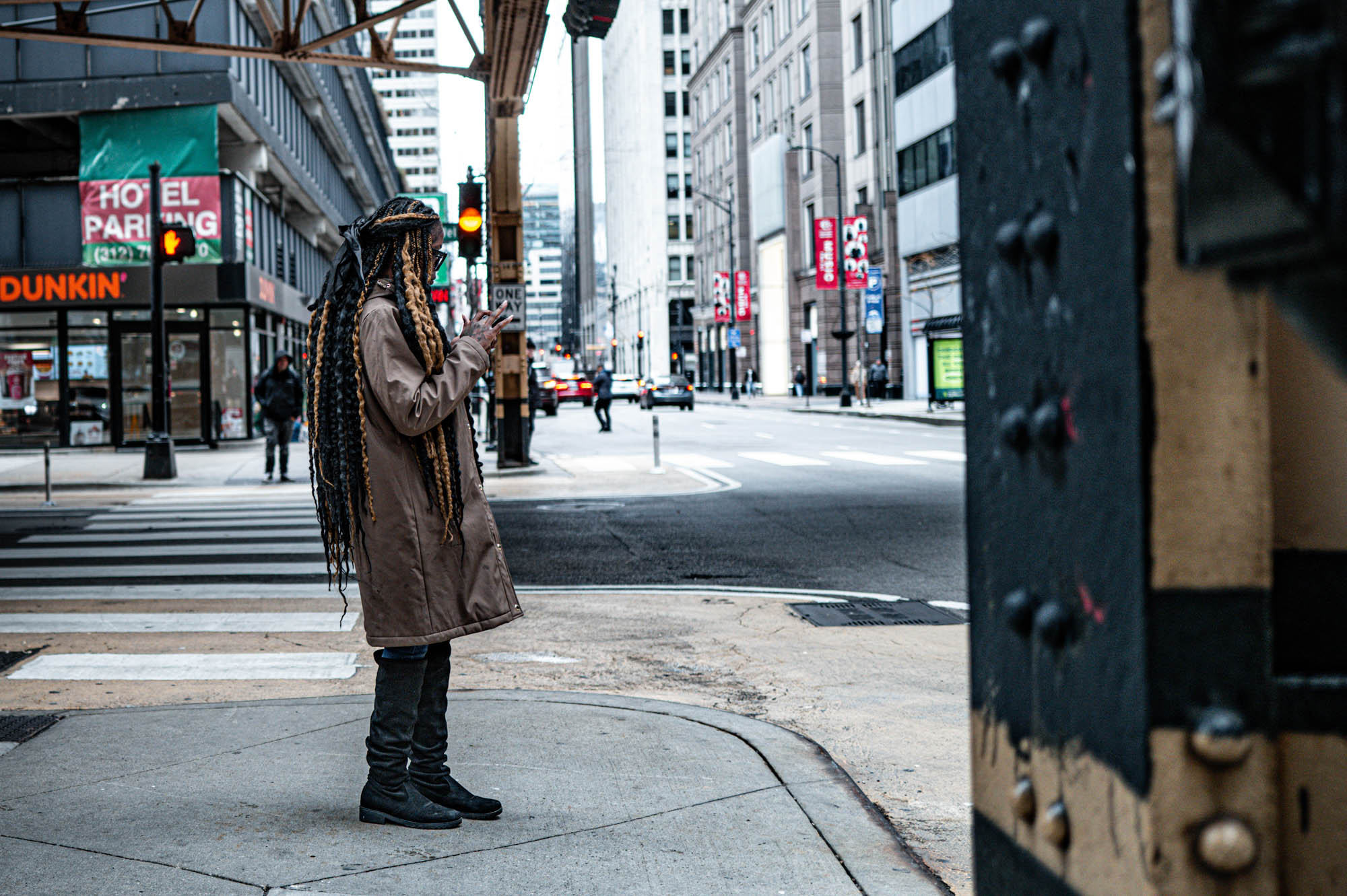Person in coat stands at city intersection, using phone, surrounded by tall buildings and shops.