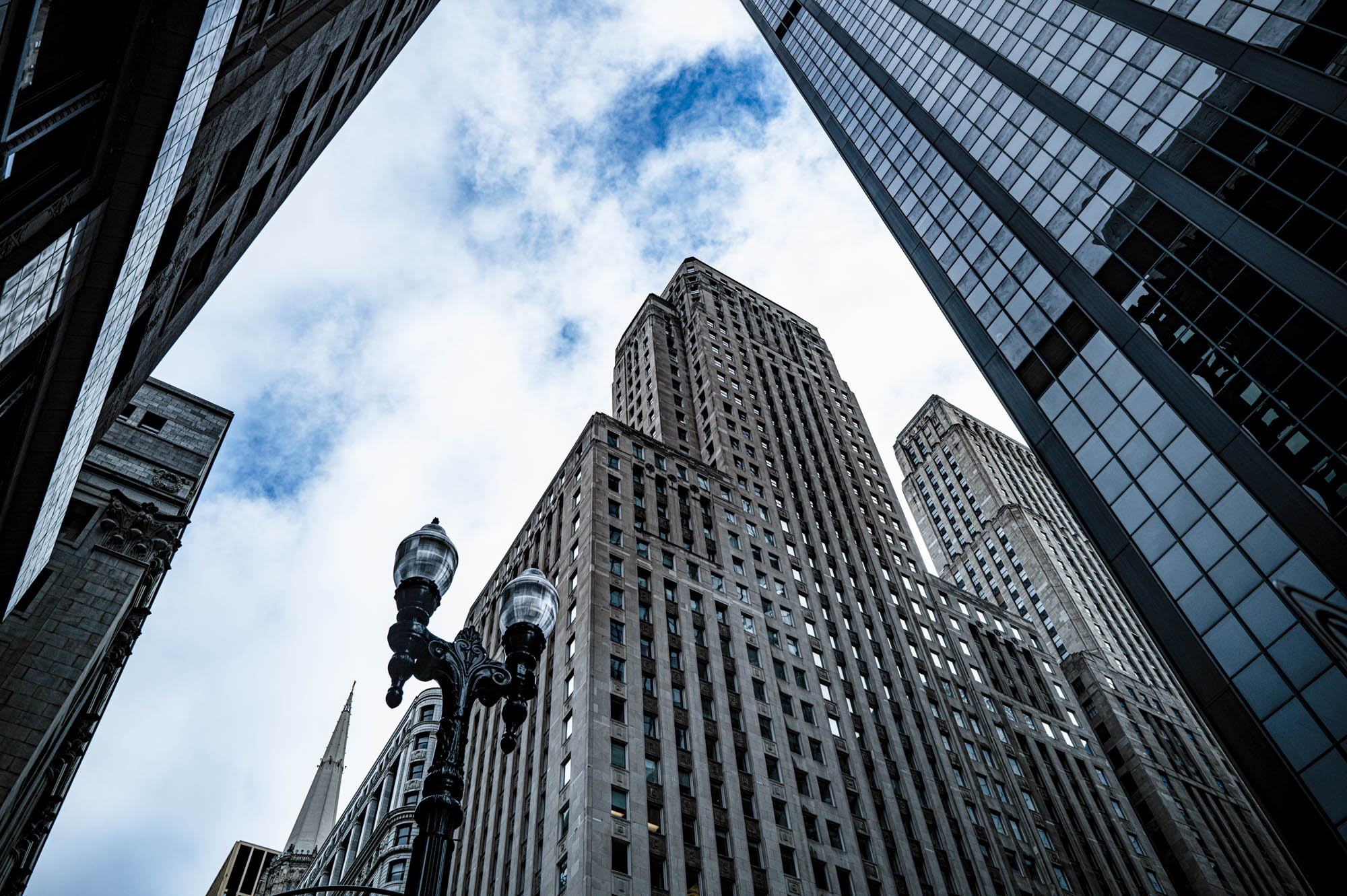 Tall city skyscrapers viewed from below against a cloudy blue sky, highlighting urban architecture and design.