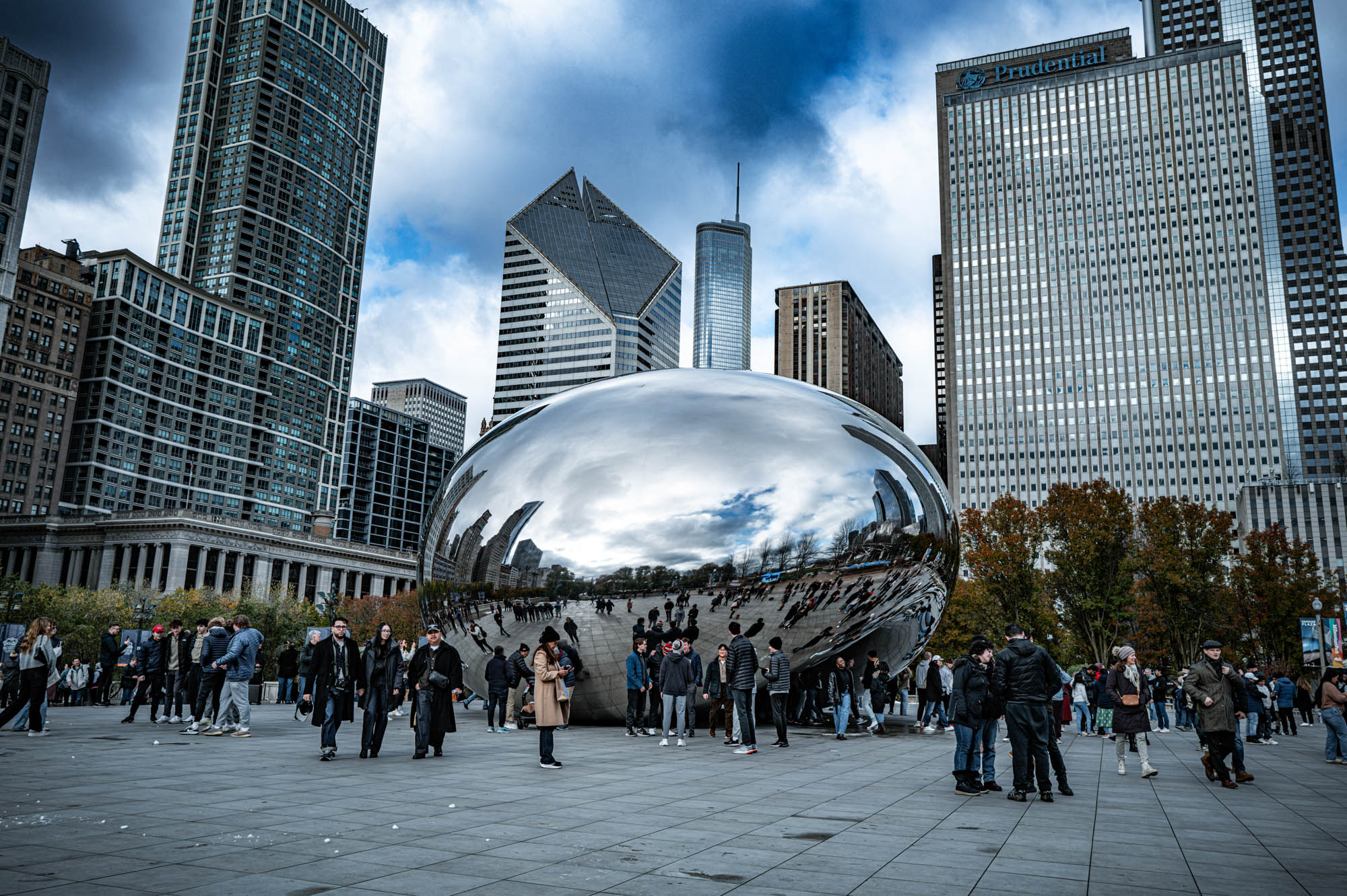 Cloud Gate in Chicago's Millennium Park, surrounded by tourists and skyscrapers under a cloudy sky.