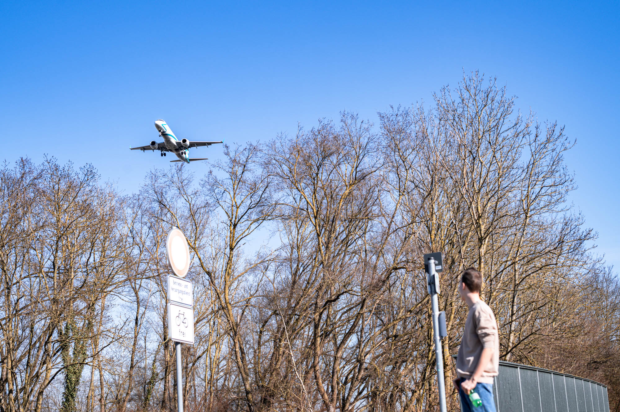 Airplane flying low over trees, with person observing from below on a sunny day, blue sky in the background.