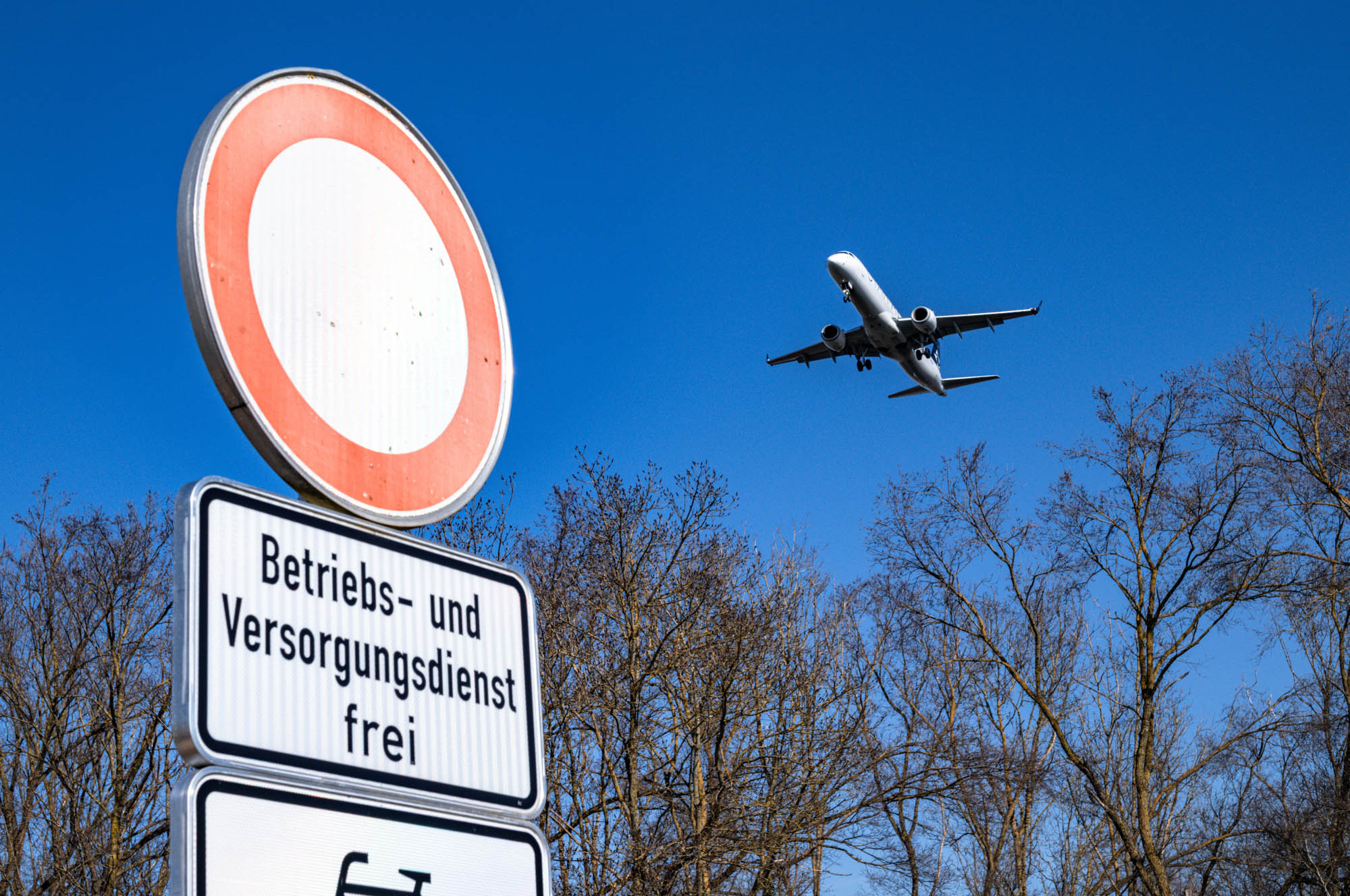 Airplane flying over trees near a Betriebs- und Versorgungsdienst frei road sign against blue sky.