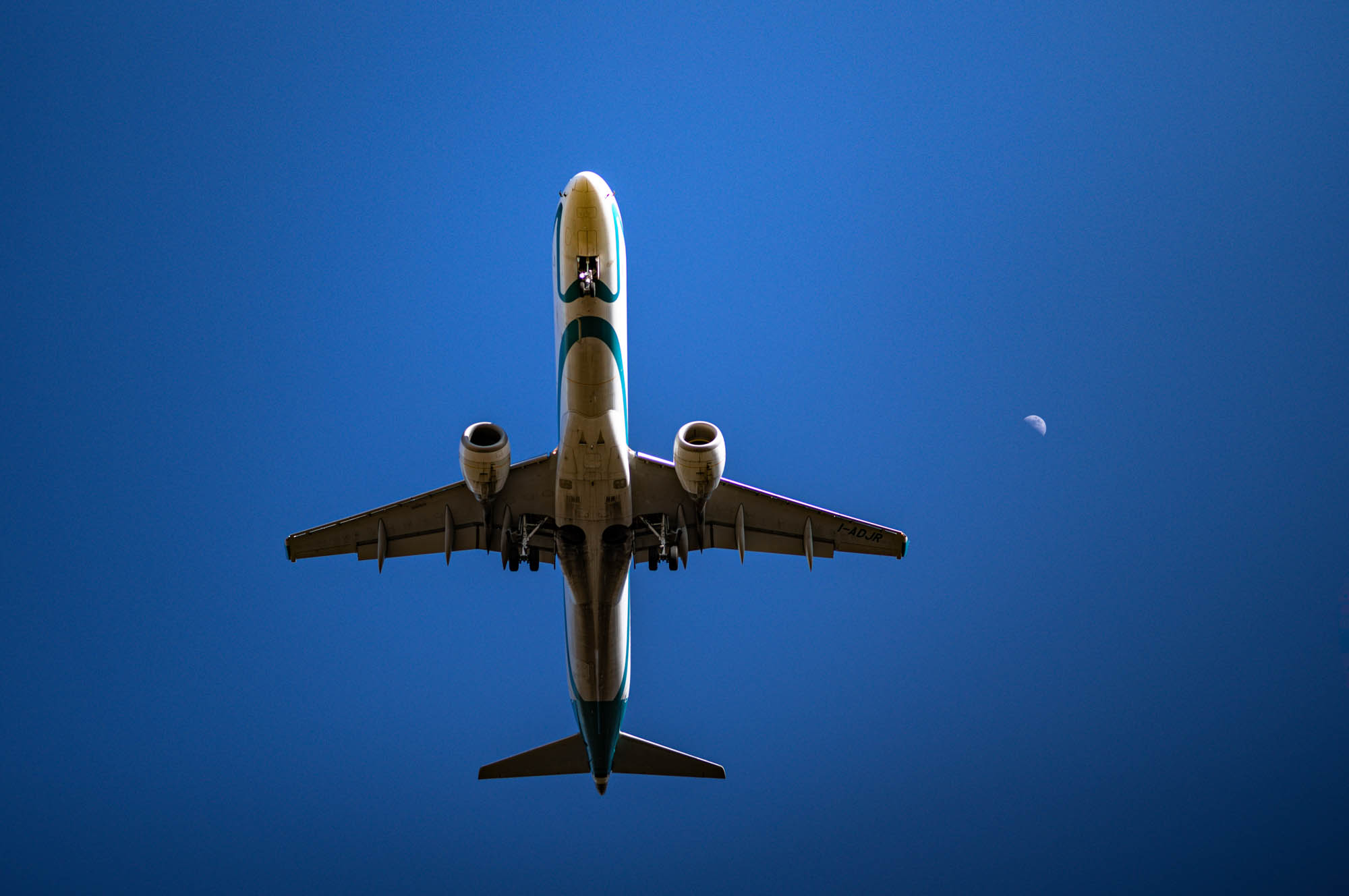 Airplane flying overhead against a clear blue sky with the moon visible in the background.