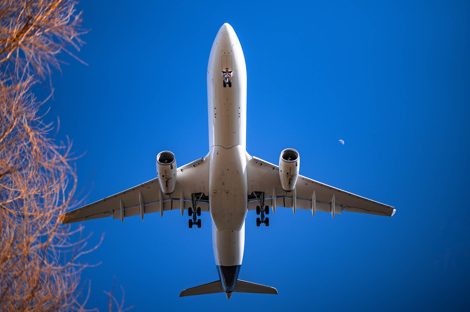 Commercial airplane flying overhead against clear blue sky with visible moon.