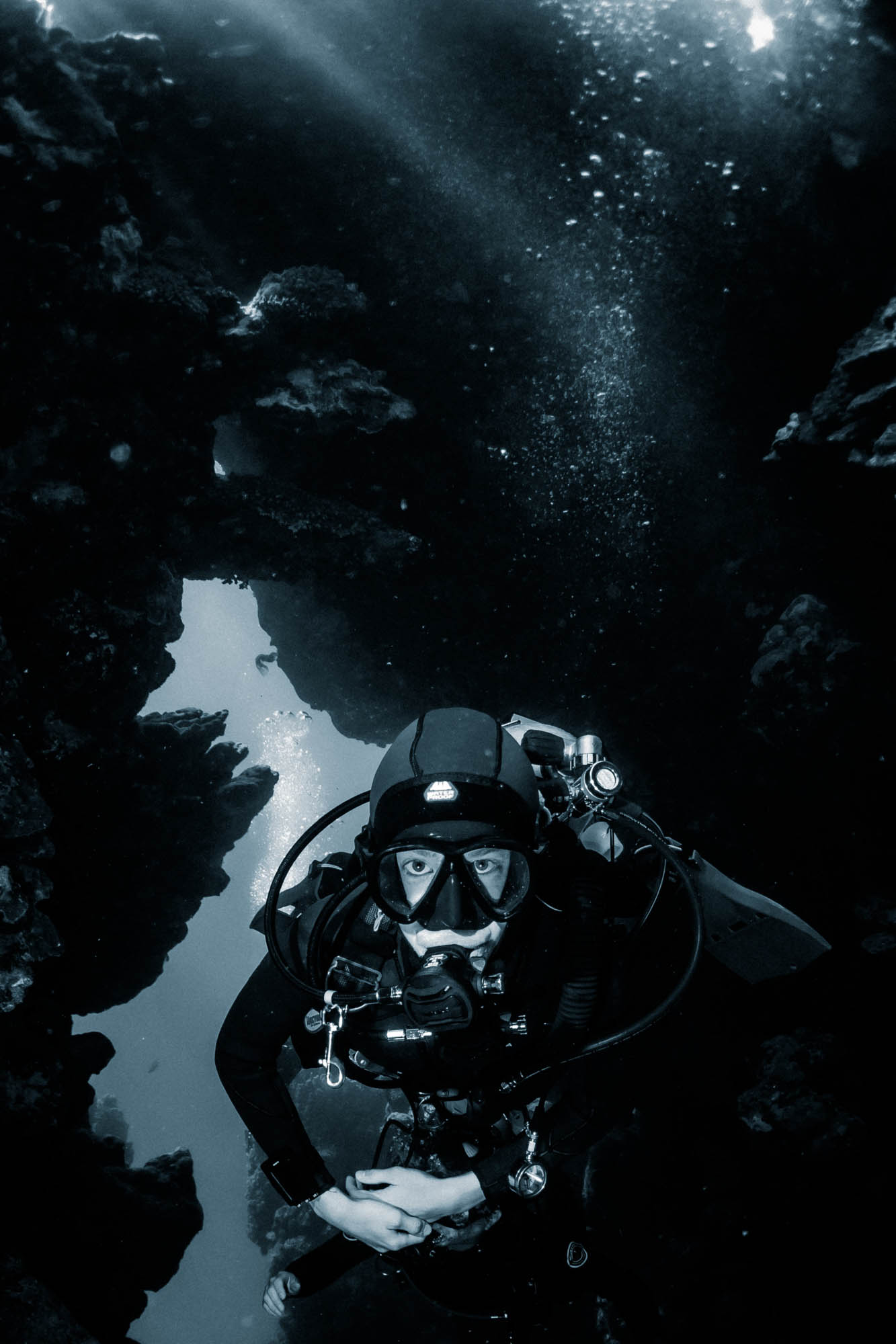 Scuba diver exploring underwater cave, surrounded by rocks and sunlight filtering through the water.