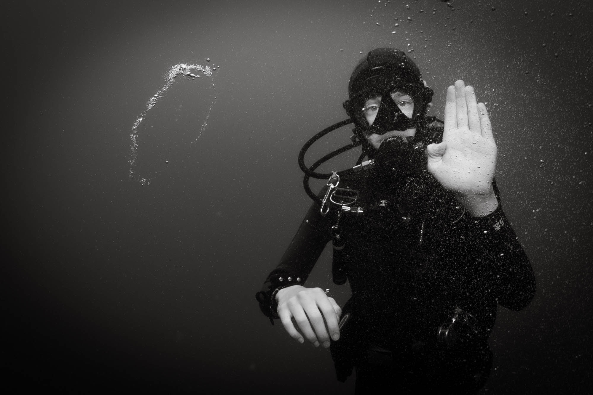 Underwater diver signals with hand in black and white photo, surrounded by bubbles.