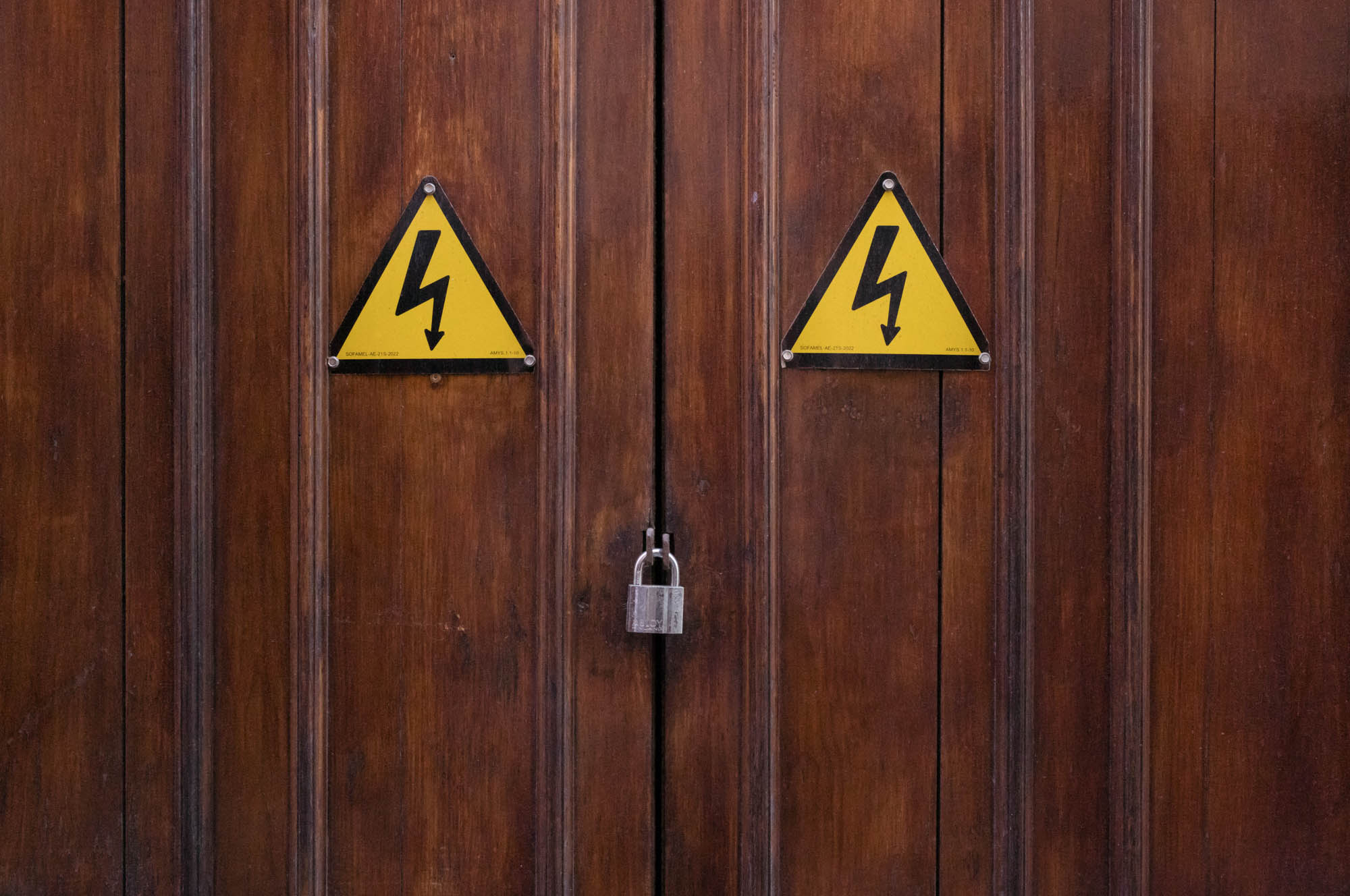 Wooden door with two high voltage warning signs and a padlock, highlighting electrical safety precautions.