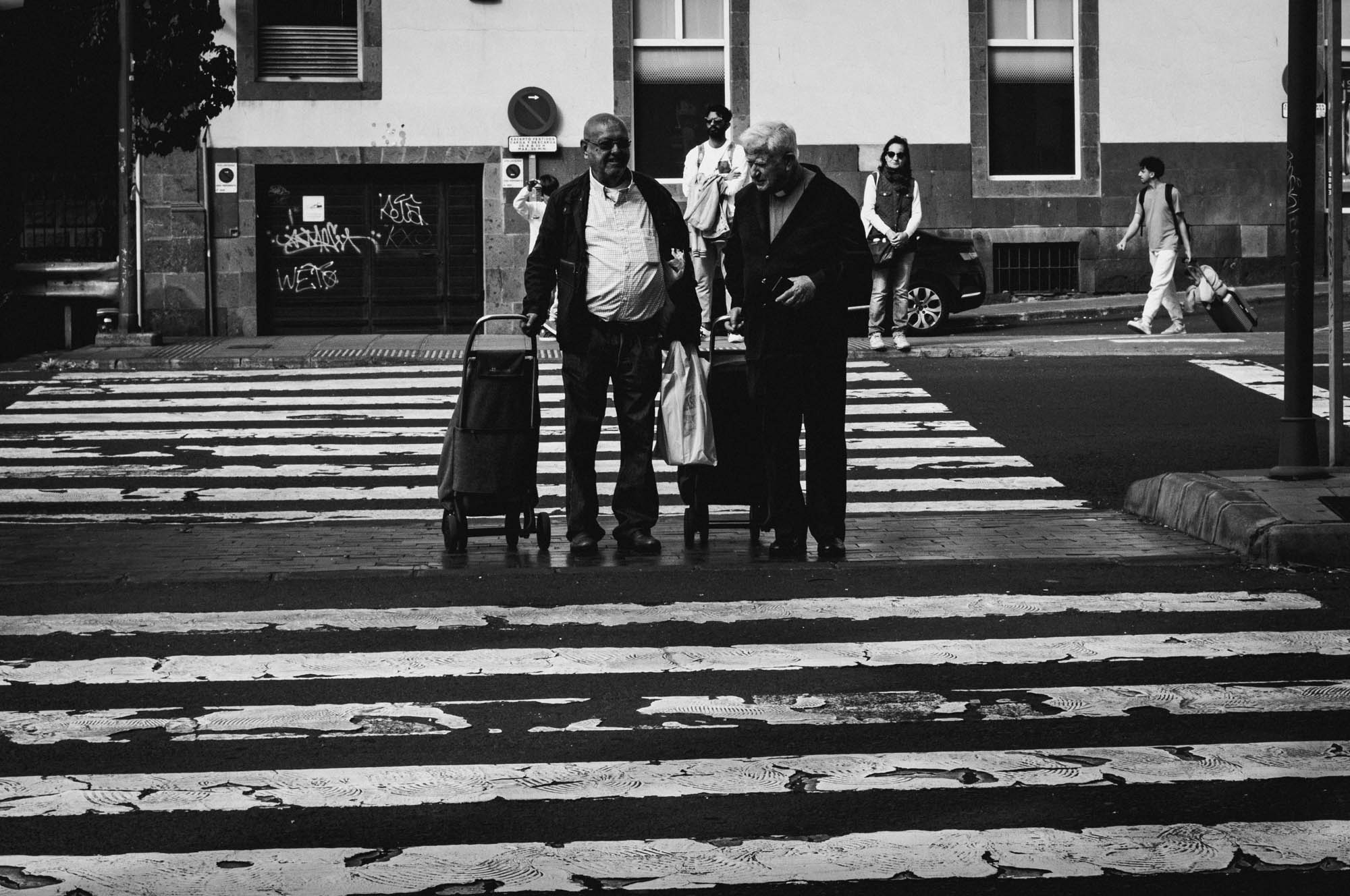 Elderly pair with carts at a crosswalk, urban setting, black and white street photography.