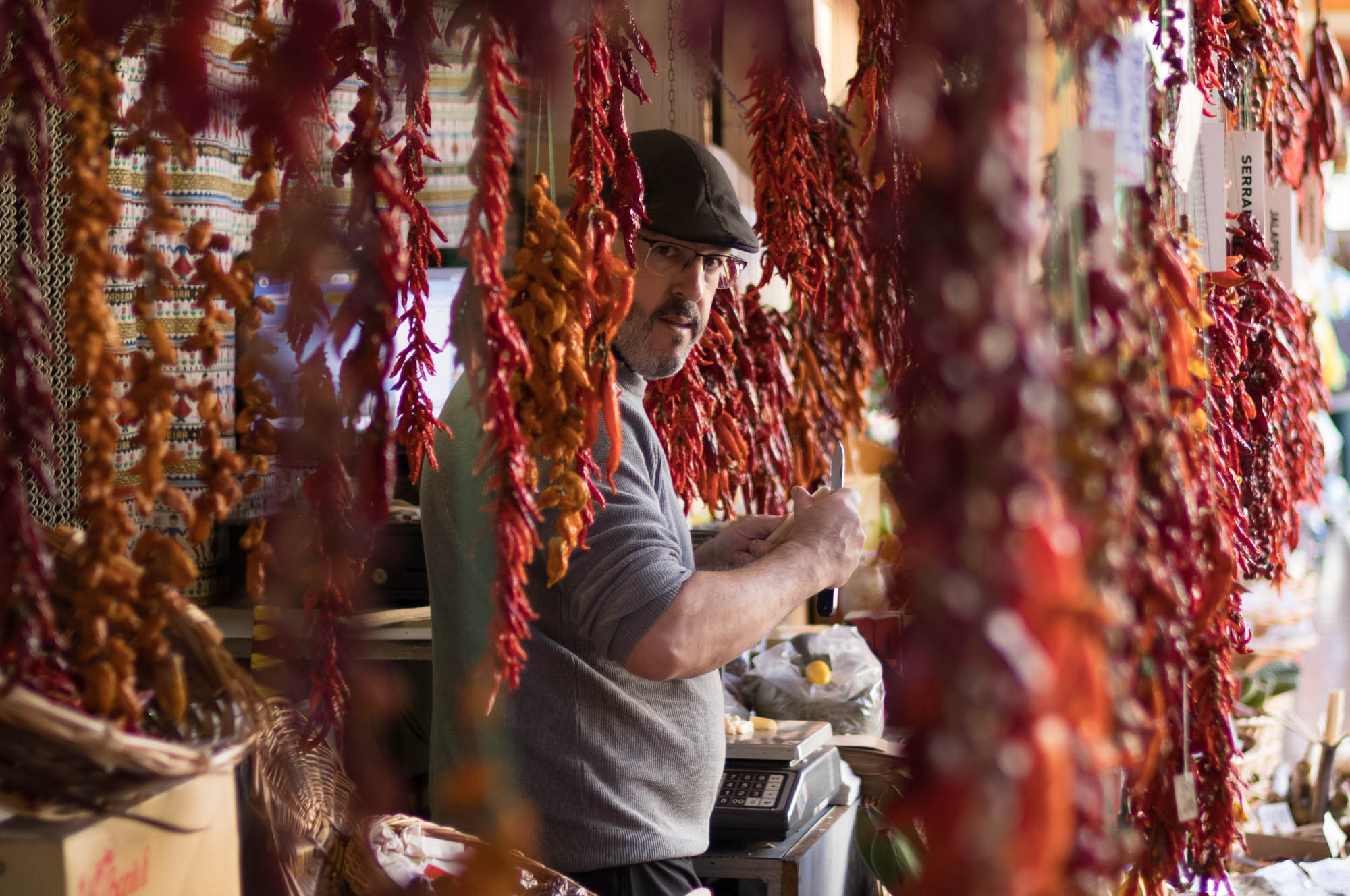Man in market stall with hanging chili peppers, preparing food.