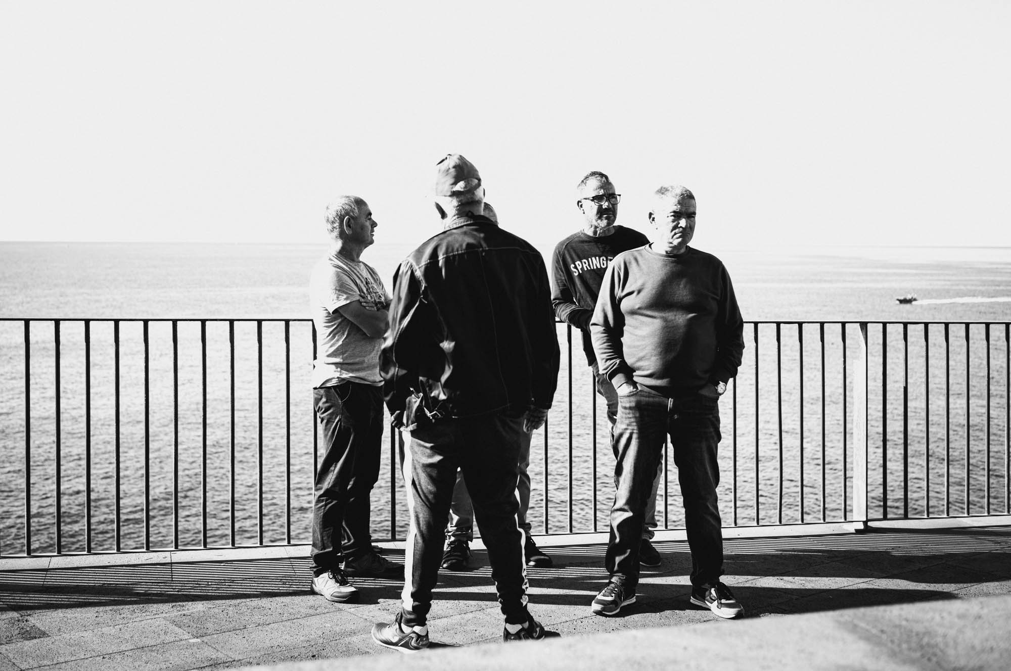 Four men standing on a seaside balcony, engaging in conversation, with an ocean view in black and white.