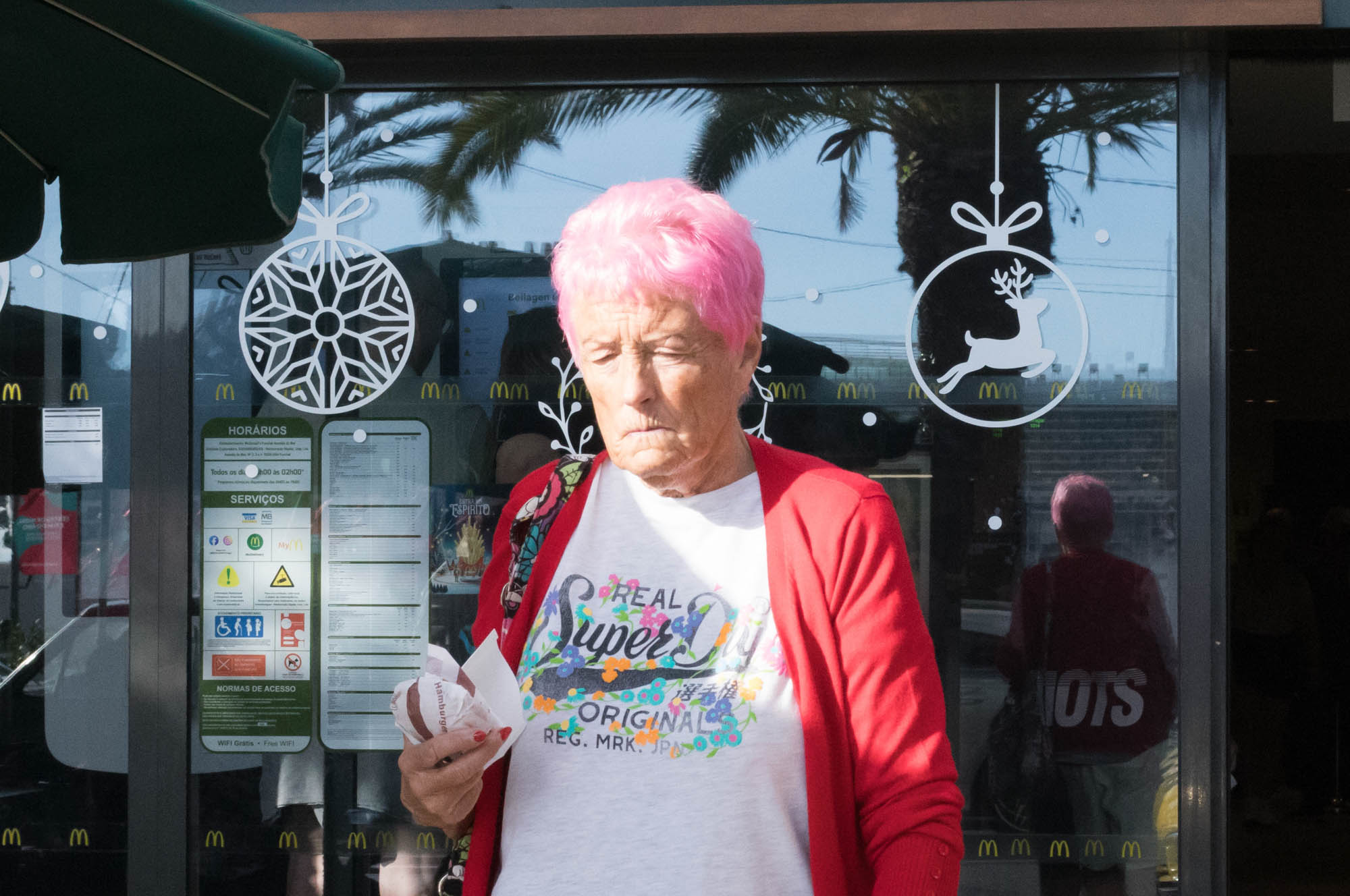 Elderly woman with pink hair and red cardigan outside a cafe, decorated with festive ornaments.