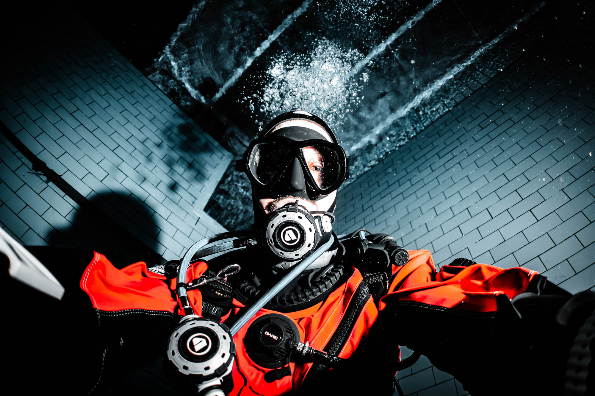 Diver in a red dry suit and mask underwater, with bubbles rising against a tiled background.