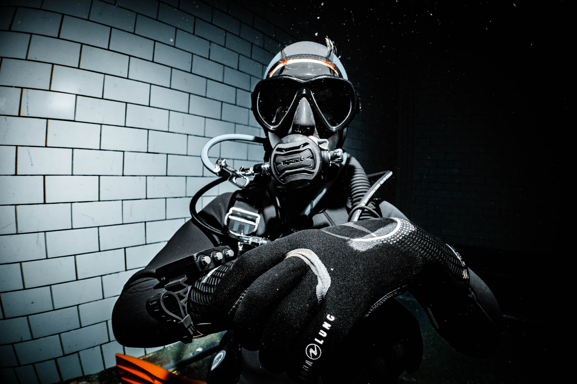 Diver in full scuba gear preparing for an underwater expedition, brick background, black wetsuit and gloves.