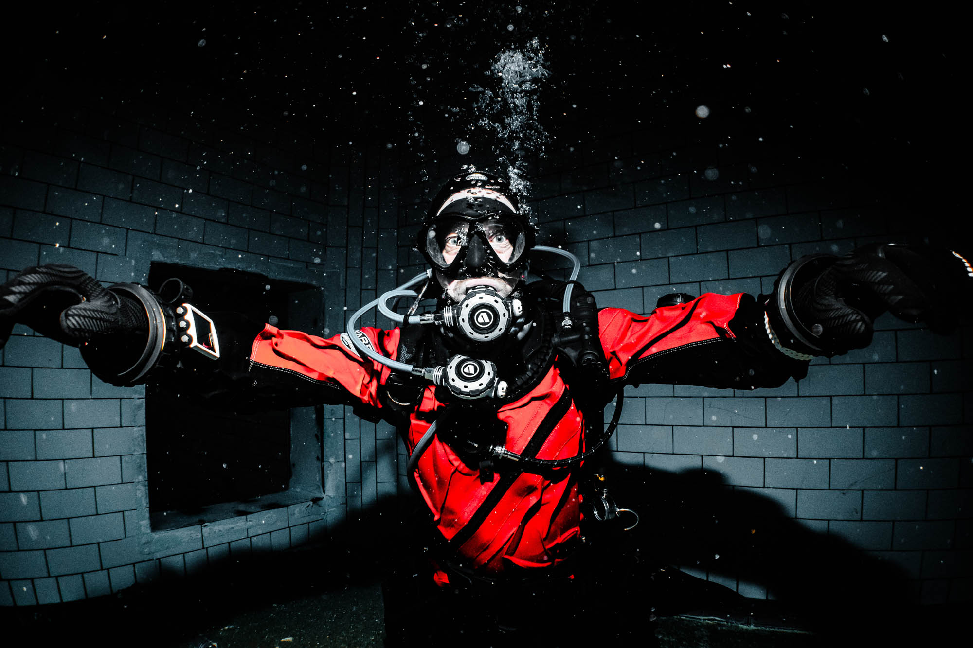 Deep-sea diver in red suit with full safety gear, underwater in dark, tiled enclosure.