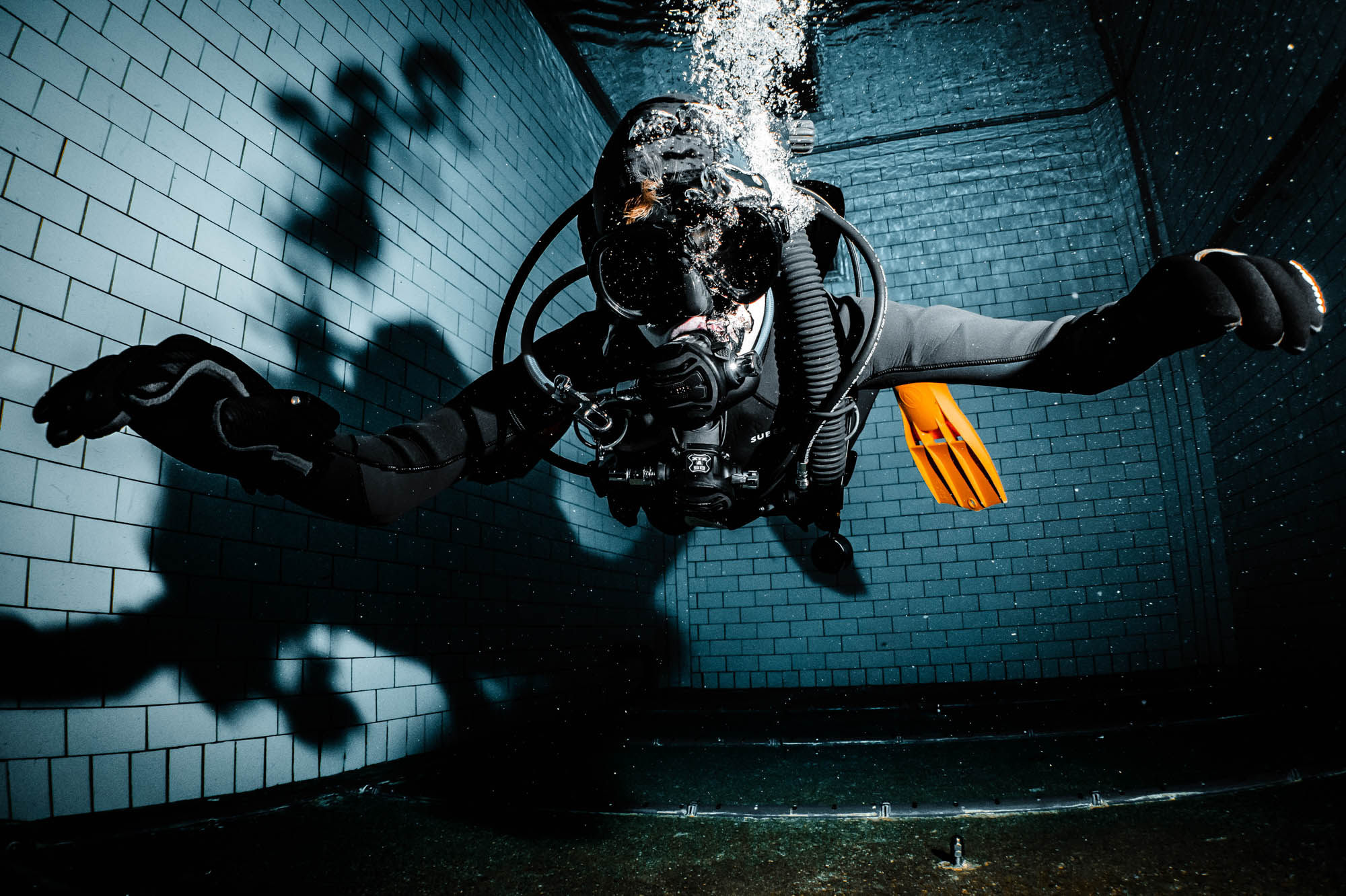 Scuba diver in black wetsuit and orange fins underwater in a tiled pool.