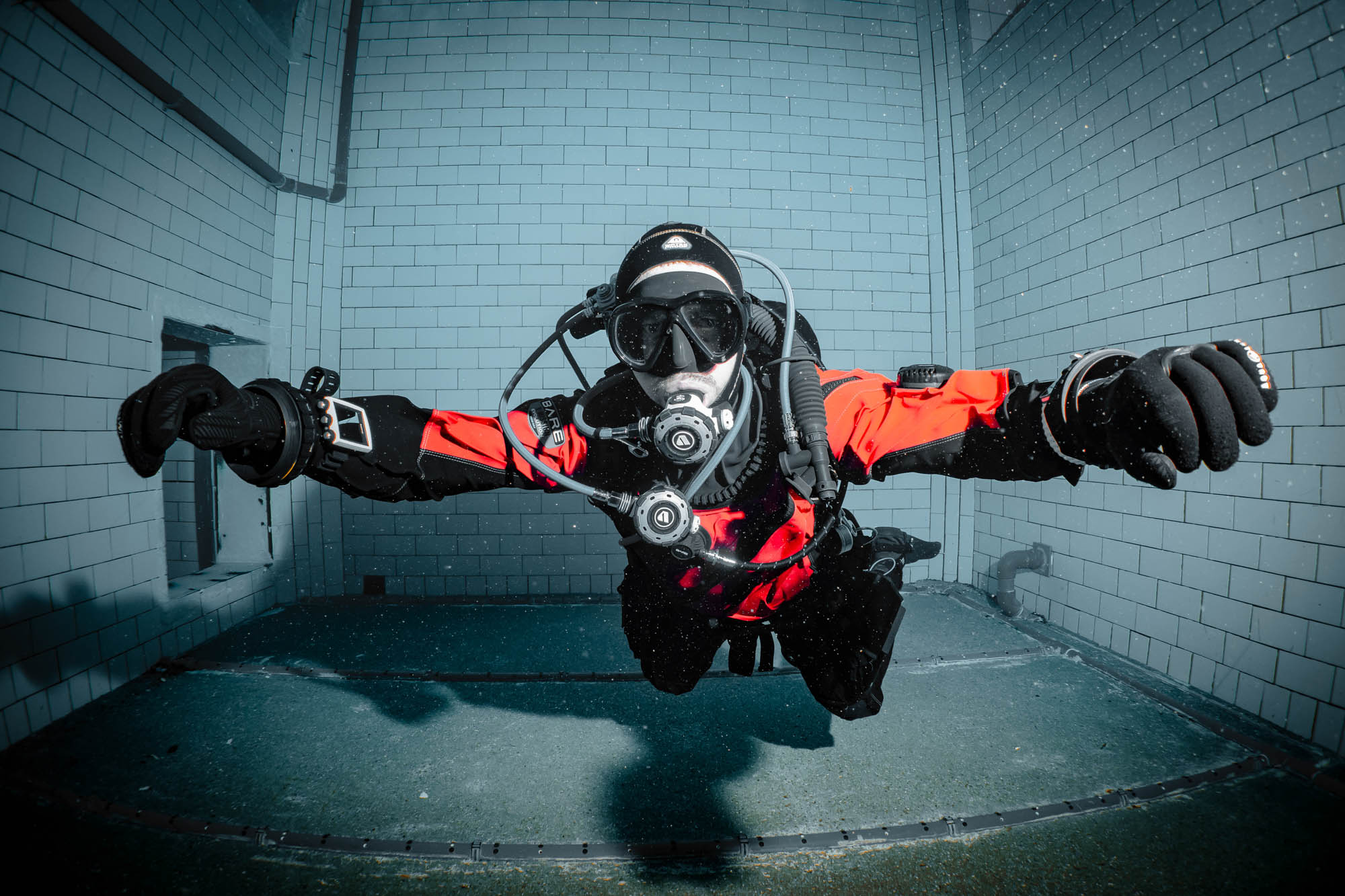 Diver in red dry suit swims underwater, wearing gear and goggles in a tiled pool.