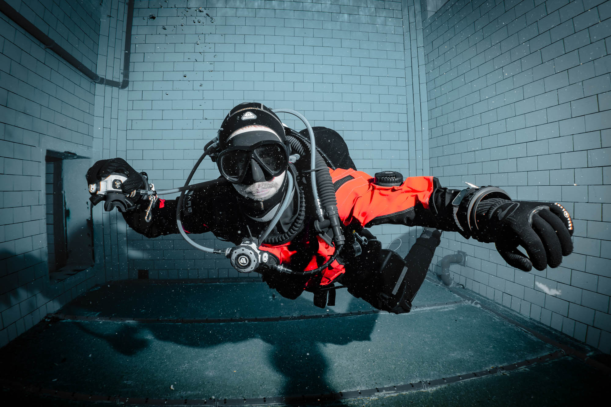 Scuba diver exploring an indoor pool with gear, wearing a red dry suit and black goggles, focused underwater training session.