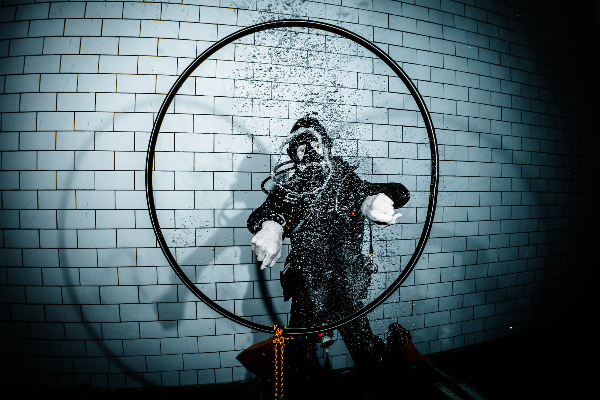 Diver in black wetsuit inside big ring, surrounded by bubbles, against a tiled underwater backdrop.