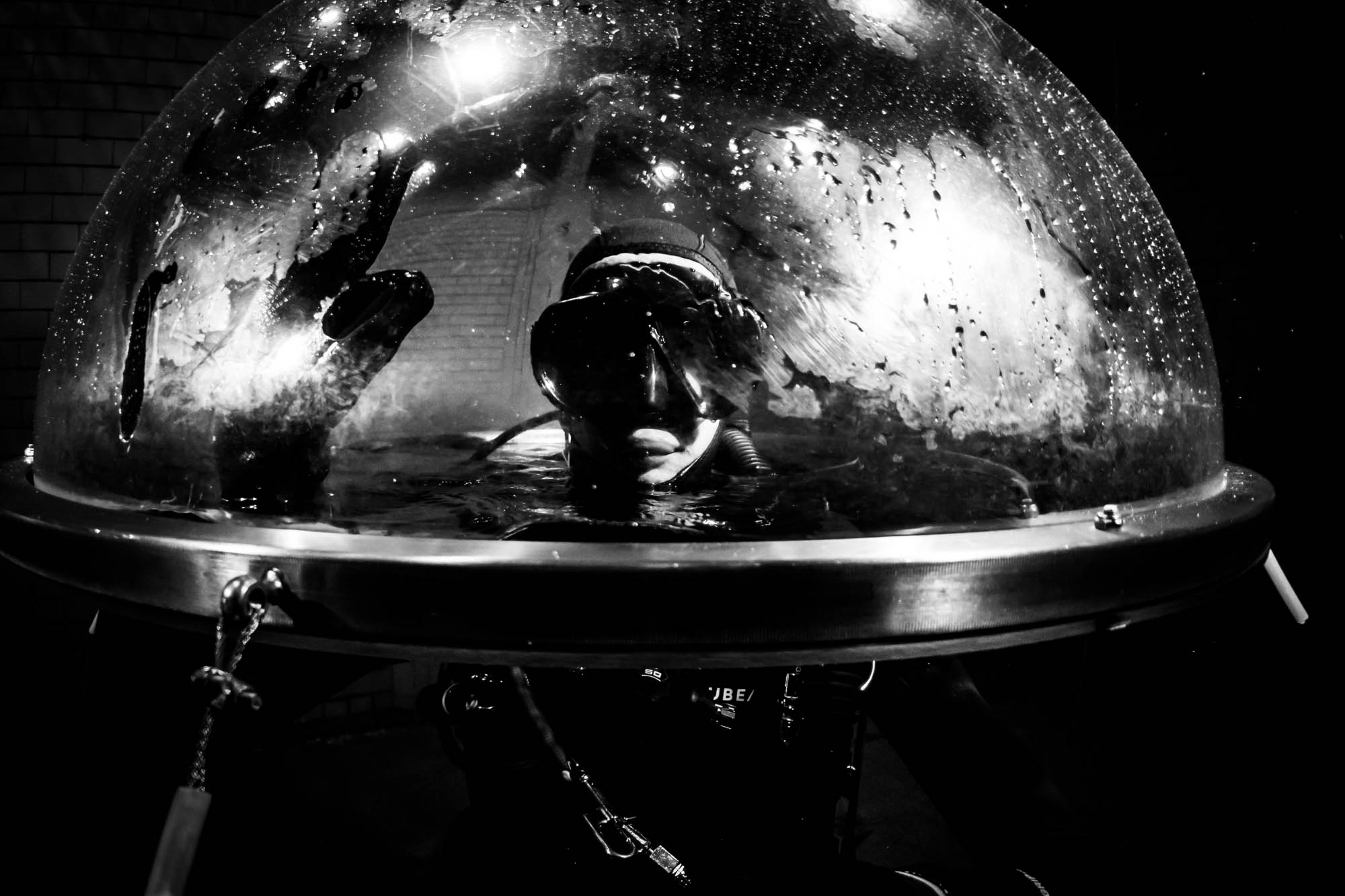 Scuba diver touching the surface of an underwater observation dome, viewed from inside, in black and white.