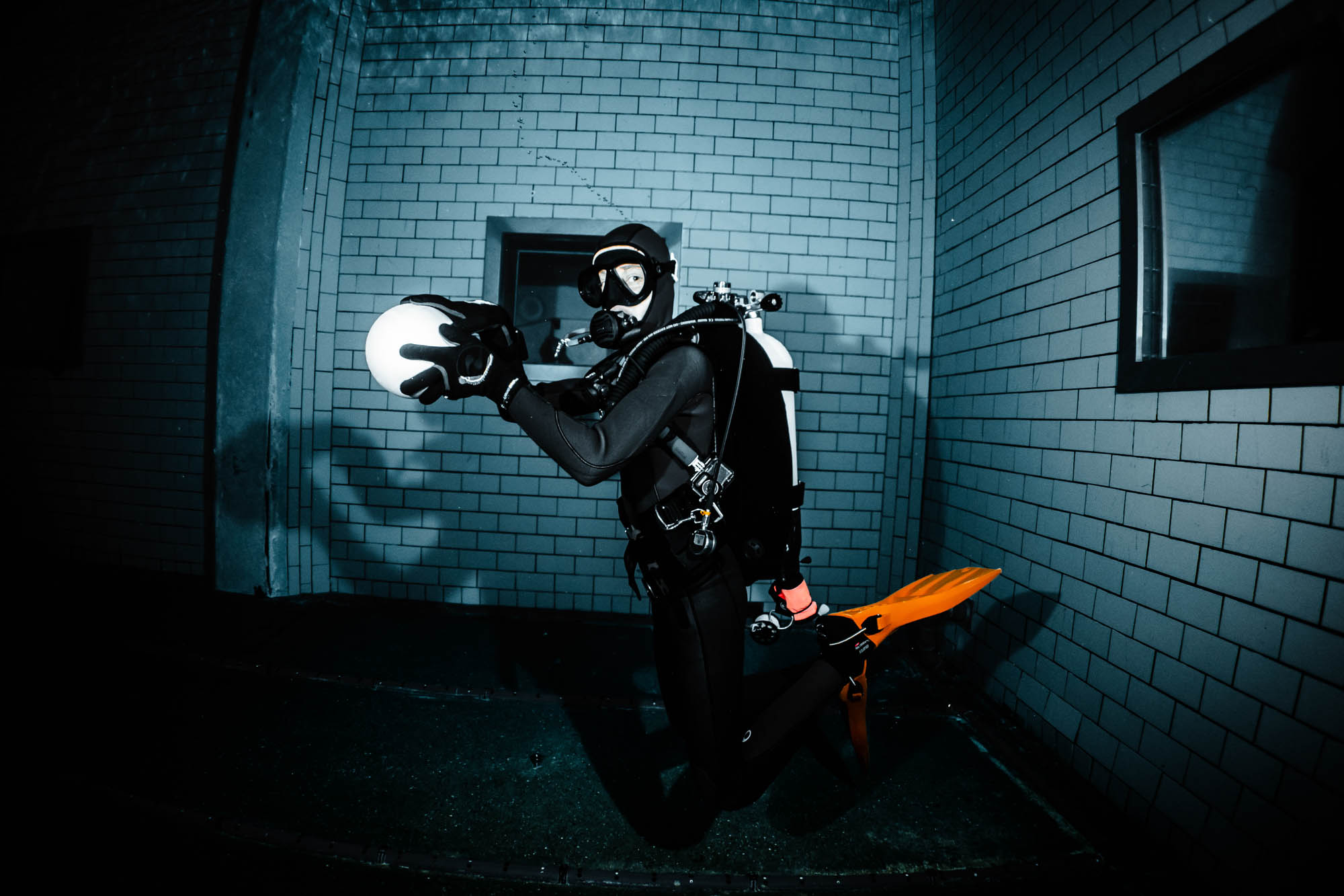 Scuba diver in full gear with orange fins exploring underwater near a brick wall, holding a white object.