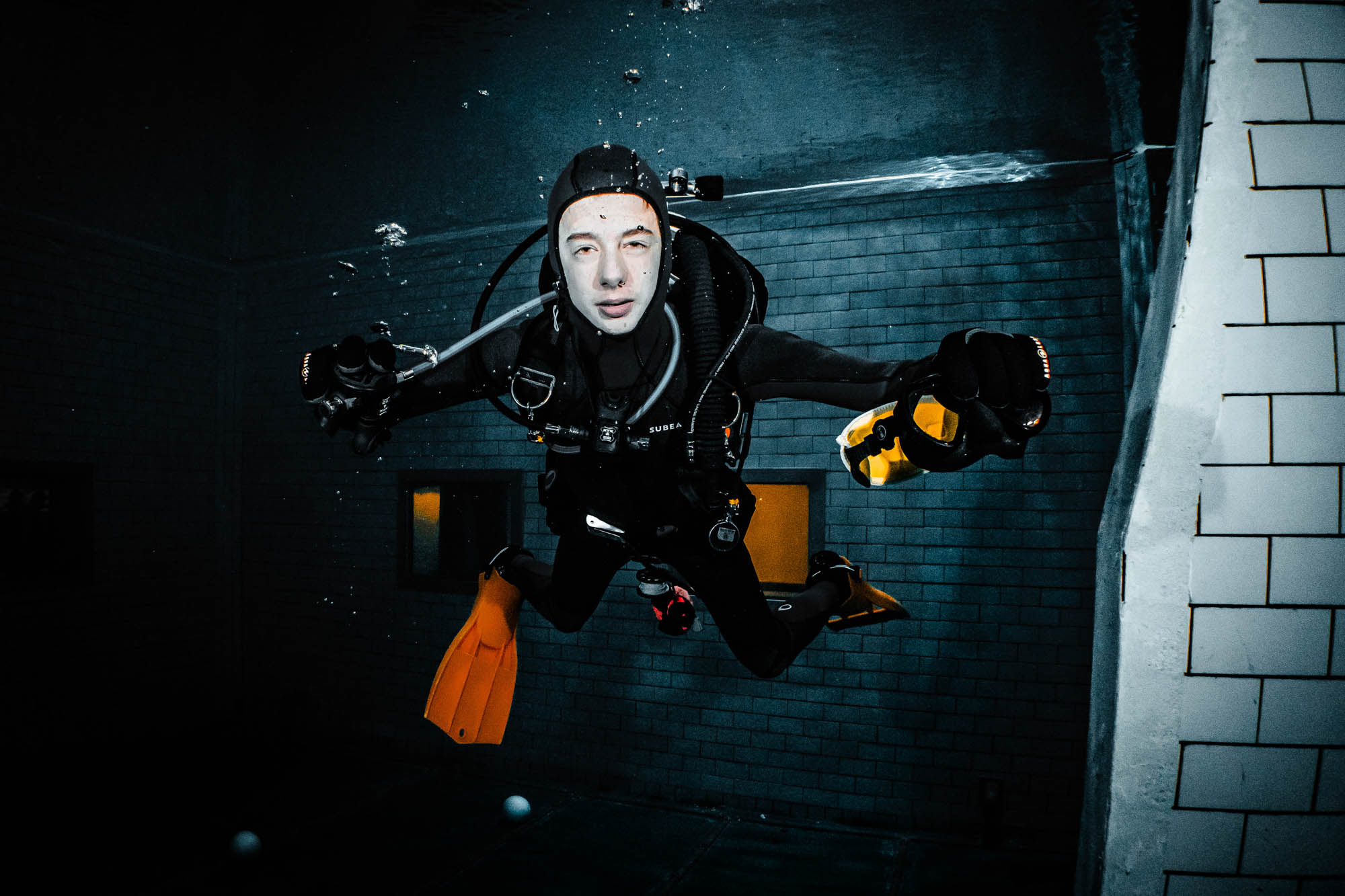 Scuba diver explores underwater with torch and gear, showcasing vibrant orange fins in a pool setting.