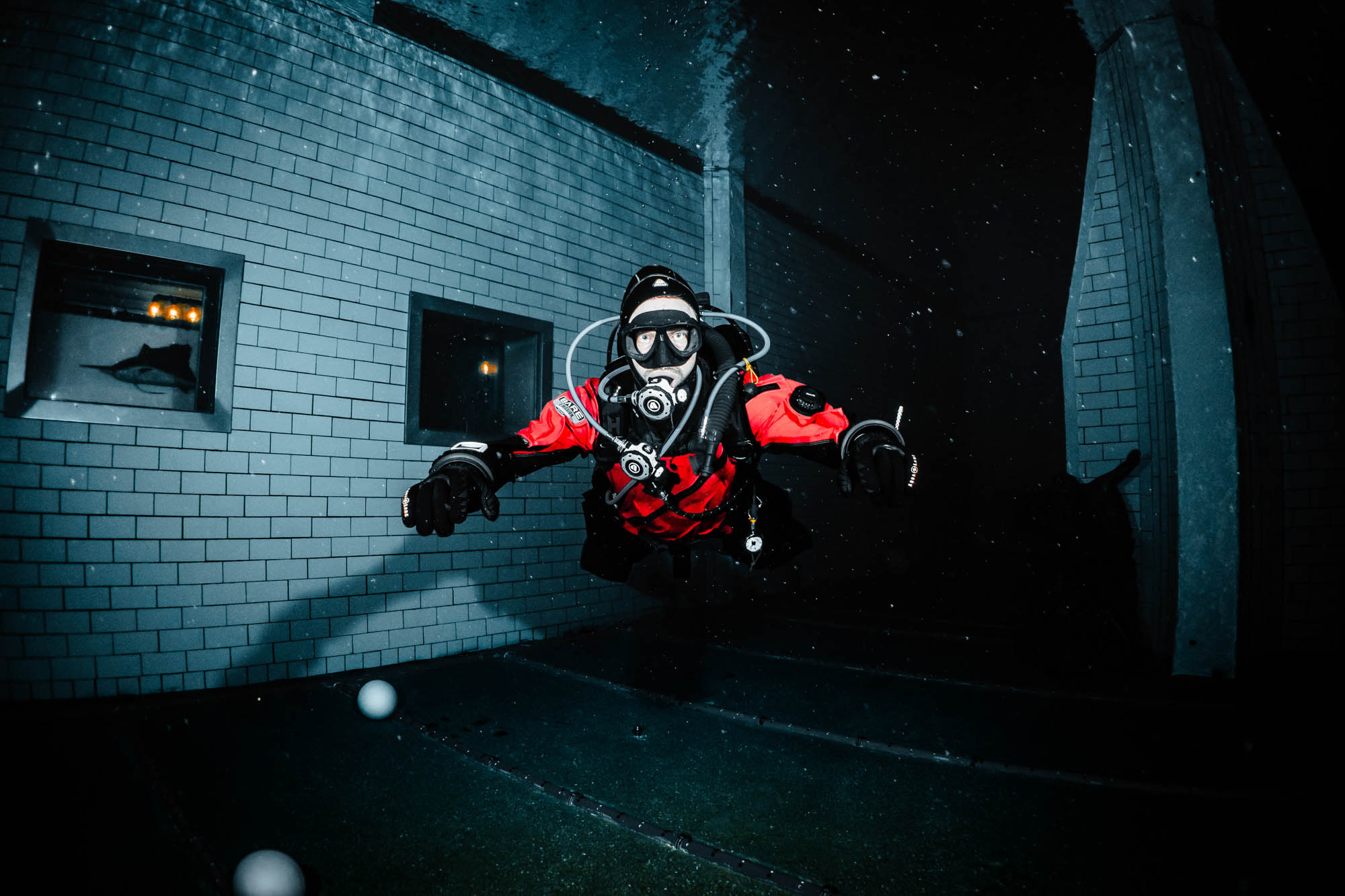 Underwater diver in red suit exploring a pool, with tiled walls and reflective windows in the background.