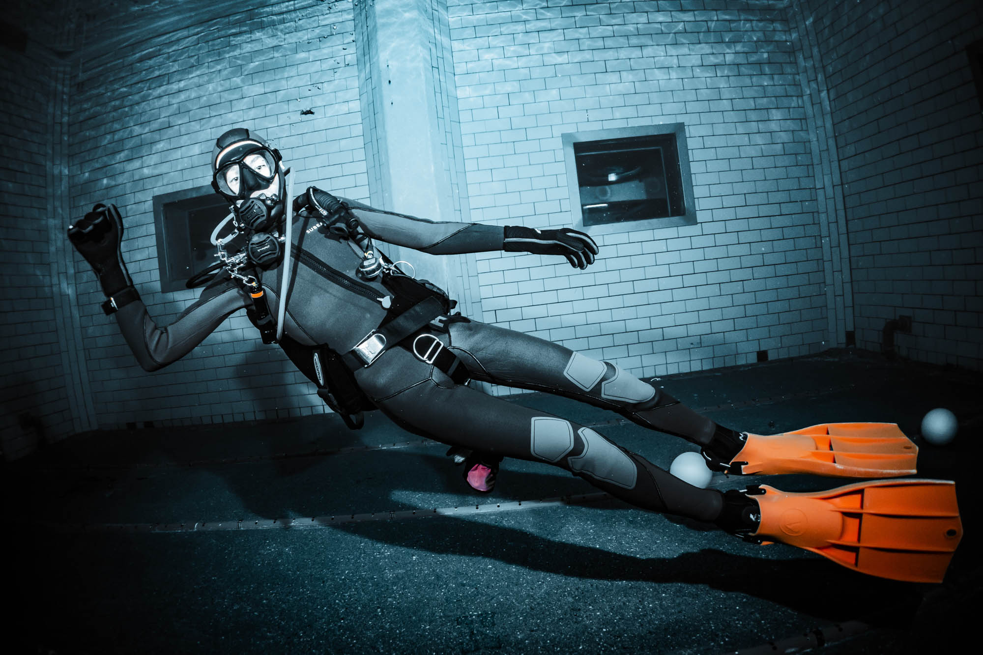 Scuba diver in full gear explores underwater with orange fins, brick wall background, in blue-toned lighting.