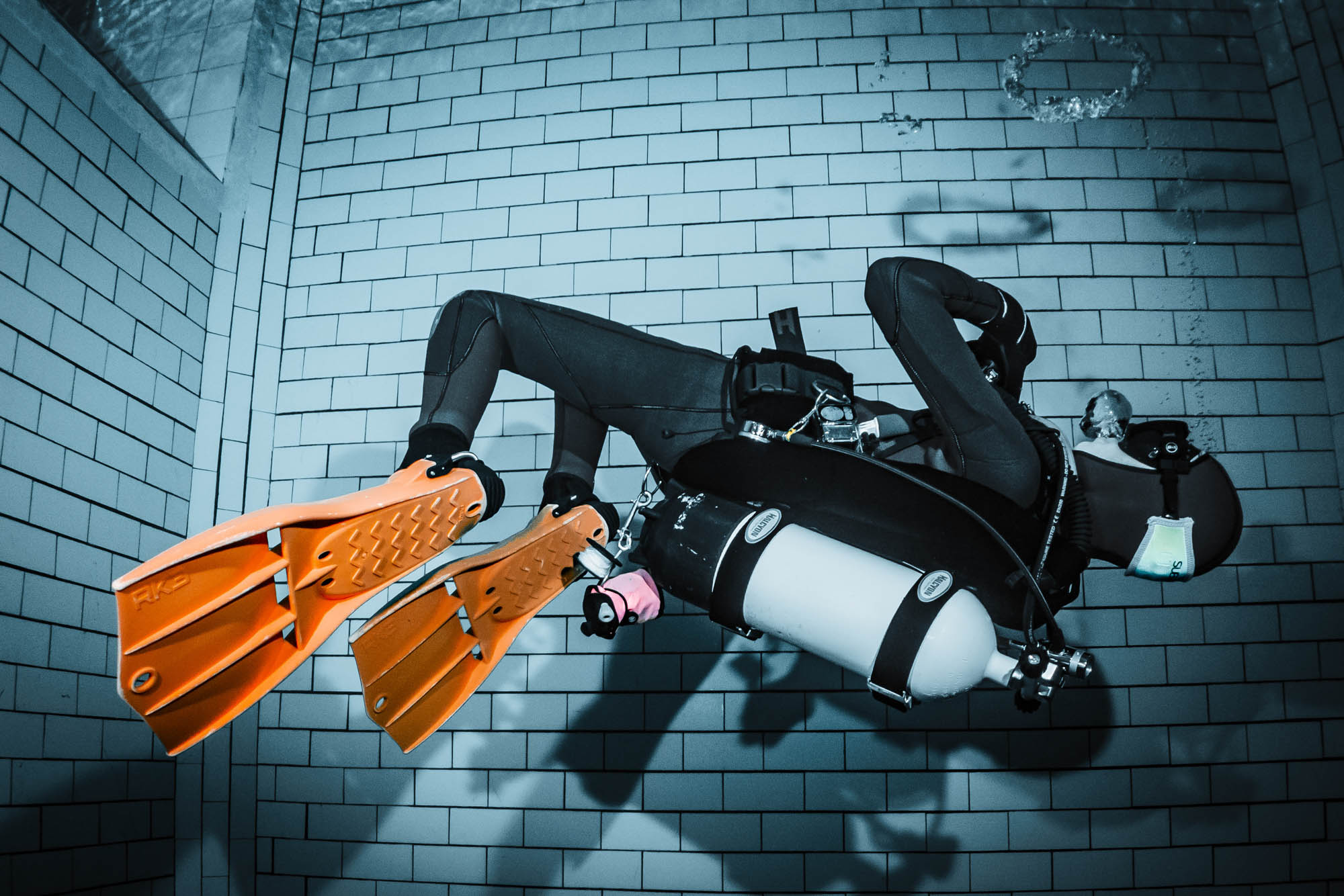 Scuba diver in gear with orange fins swims underwater alongside a tiled pool wall.