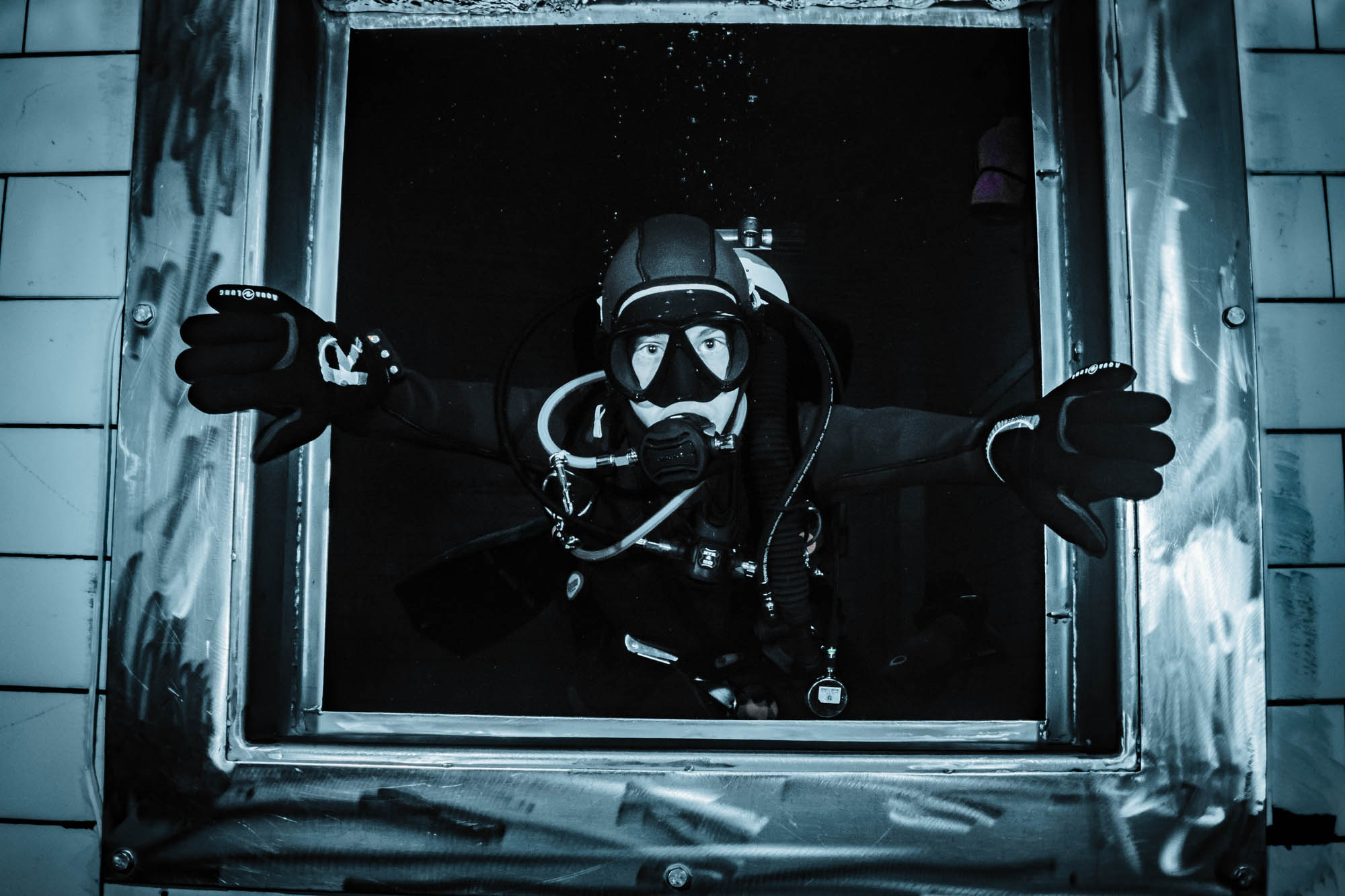 Scuba diver exploring underwater, framed by a metal window, wearing full gear and reaching out with both hands.