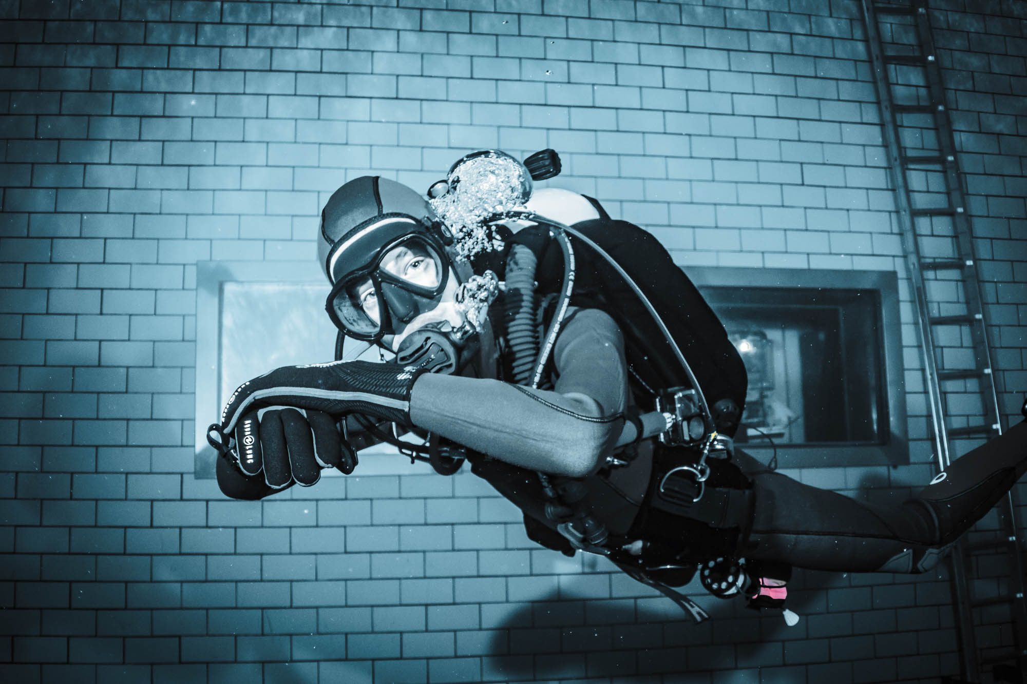 Scuba diver underwater in pool, wearing full gear, near tiled wall, creating bubbles from breathing apparatus.