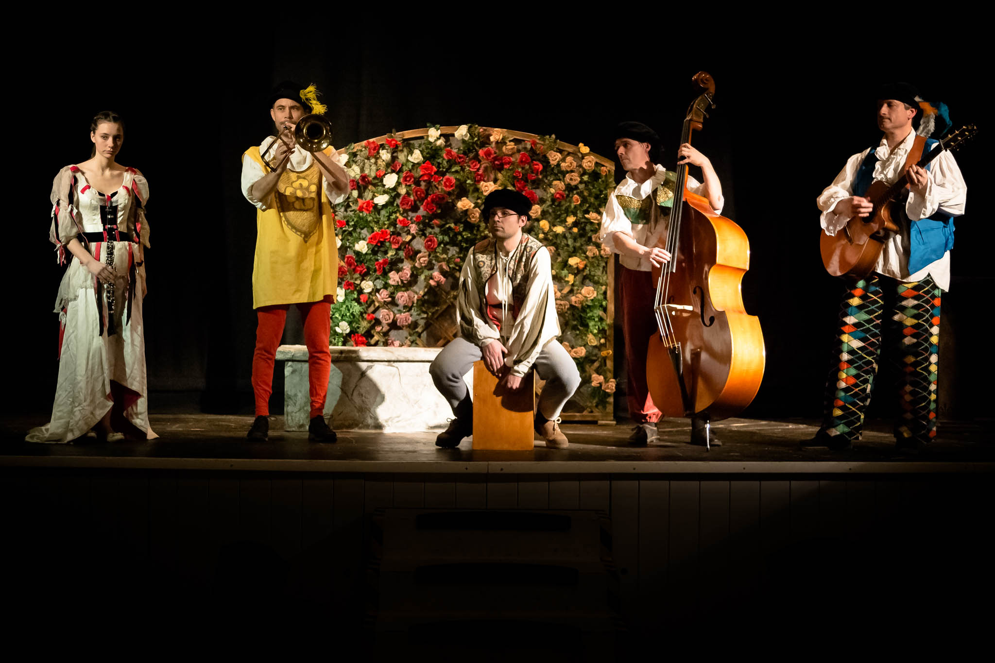 Performers in colorful medieval costumes with instruments on stage, vibrant floral backdrop, dramatic lighting.