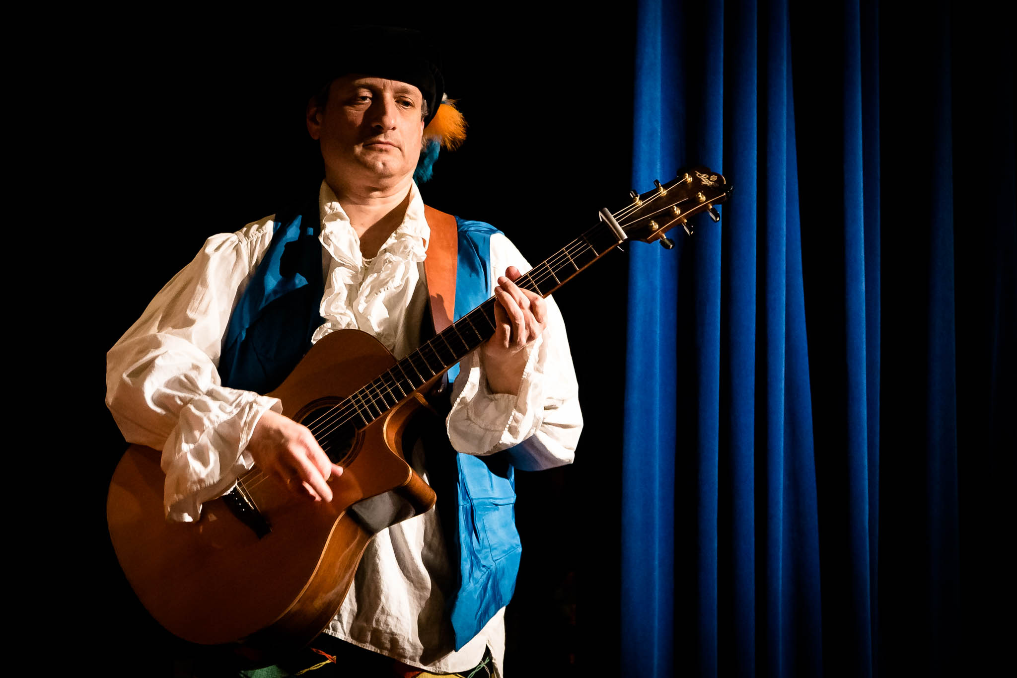 Musician in period costume playing guitar on stage with blue curtains in the background.
