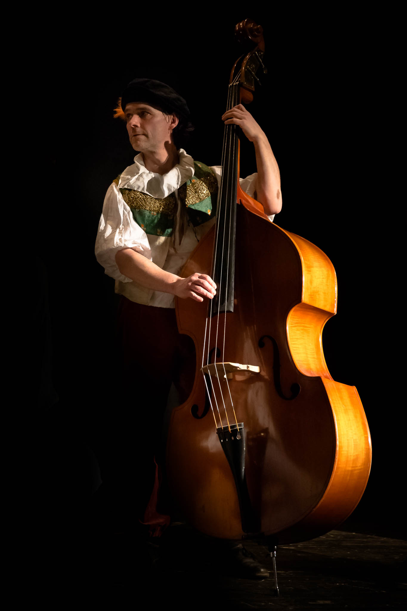 Musician in costume plays double bass on stage, highlighted by dramatic lighting against a dark background.