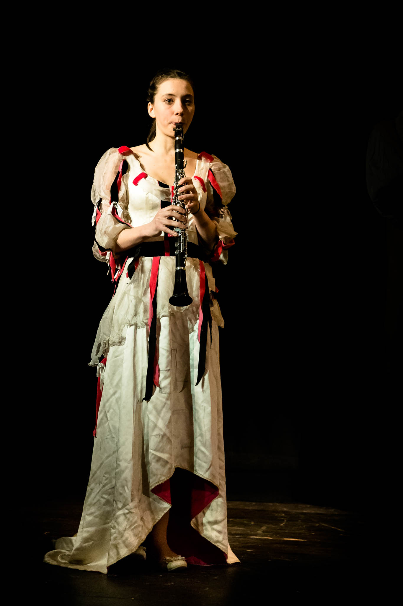 Woman in theatrical costume plays clarinet on stage, spotlight highlighting her performance, dark background.