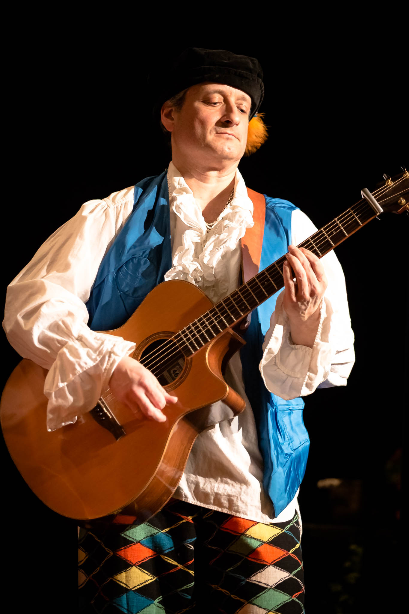 Musician in colorful costume plays acoustic guitar on stage, wearing a blue vest and feathered hat.