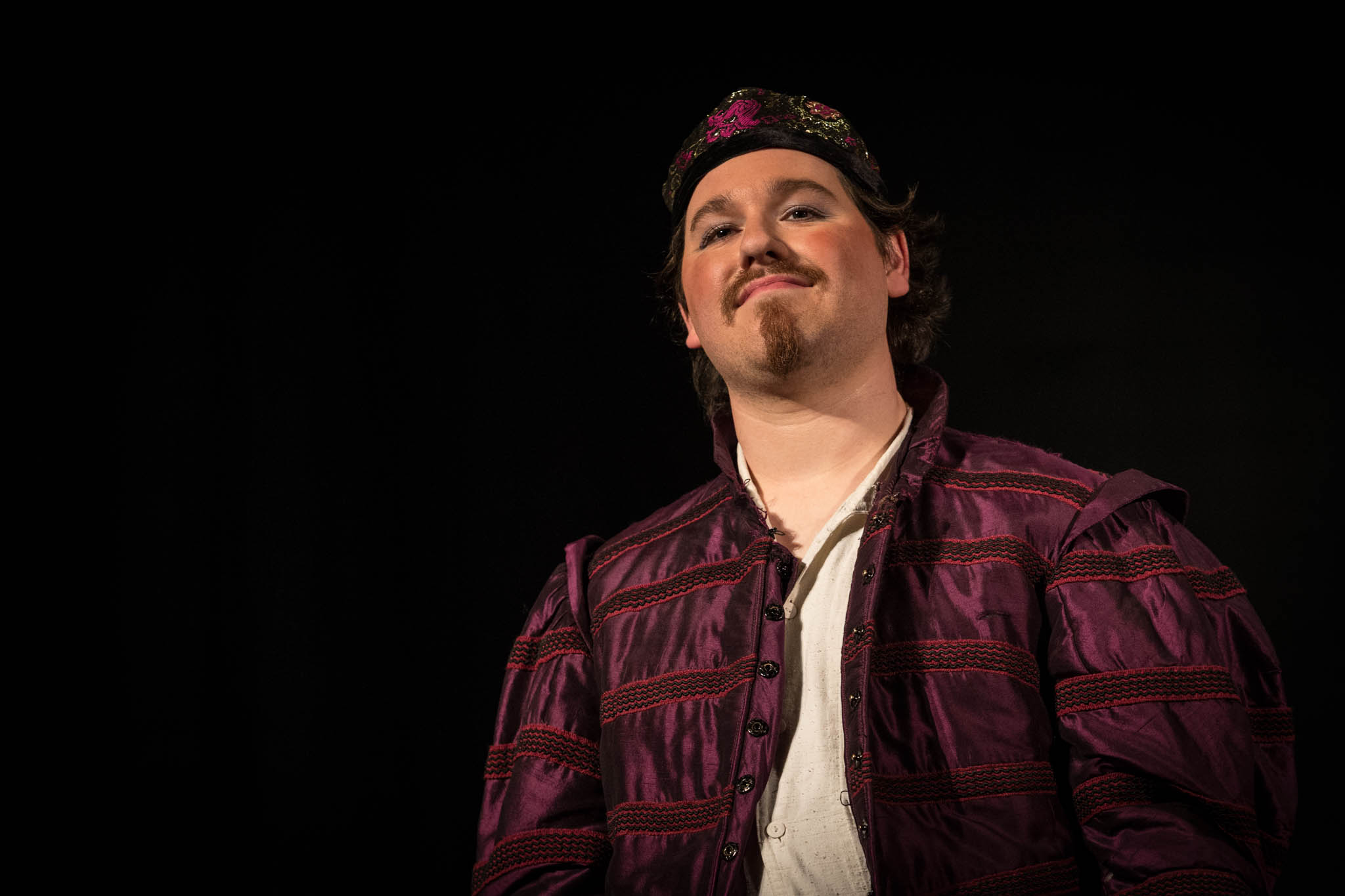 Man wearing Renaissance-style outfit and hat against dark background, posing confidently.