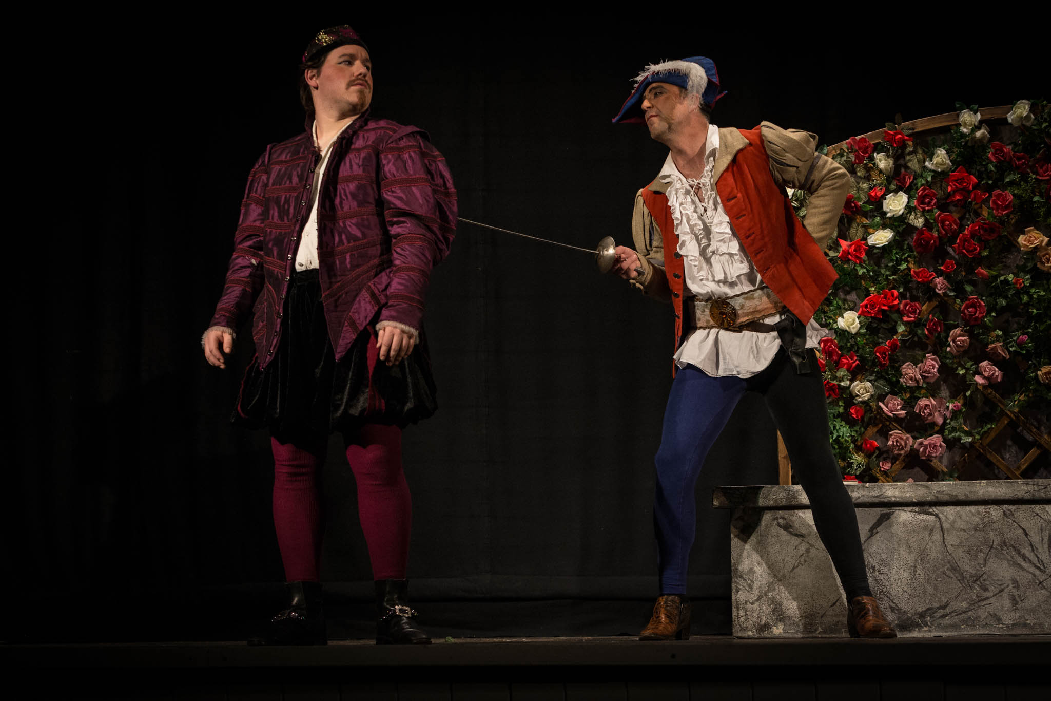 Two actors in Renaissance costumes performing a dramatic sword fight on stage with a floral backdrop.