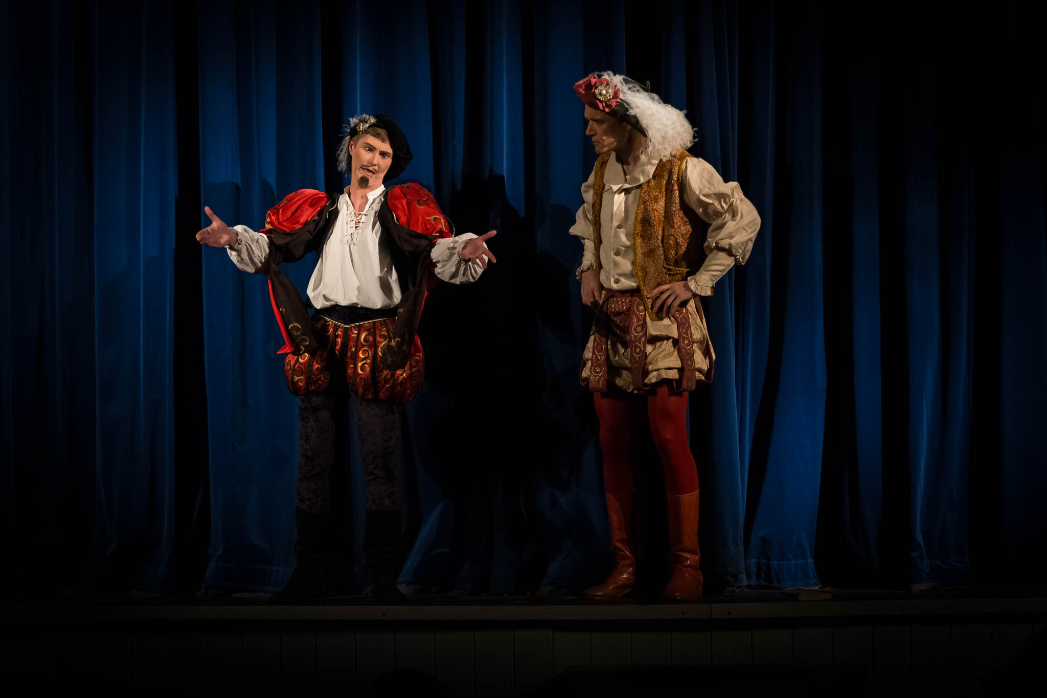 Two actors in colorful Renaissance costumes perform on stage under dramatic lighting against blue curtains.